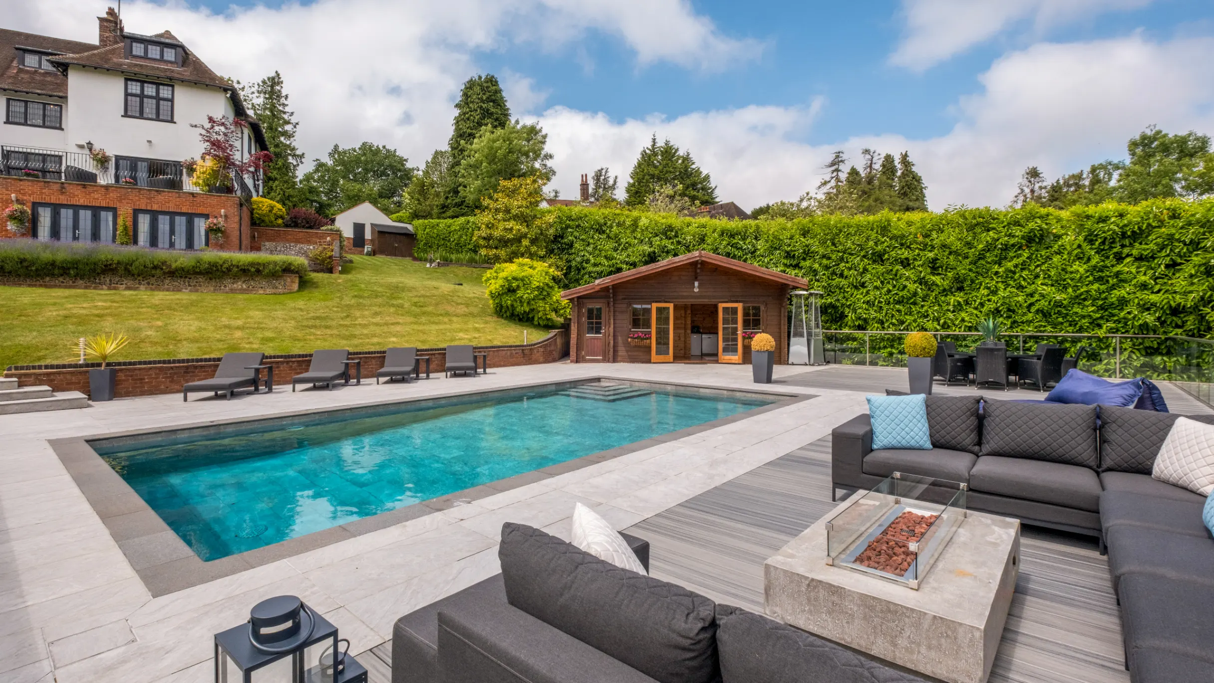 Rear garden with house exterior and lawn in the background, with a swimming pool, pool house and grey outdoor sofa with firepit in the foreground 