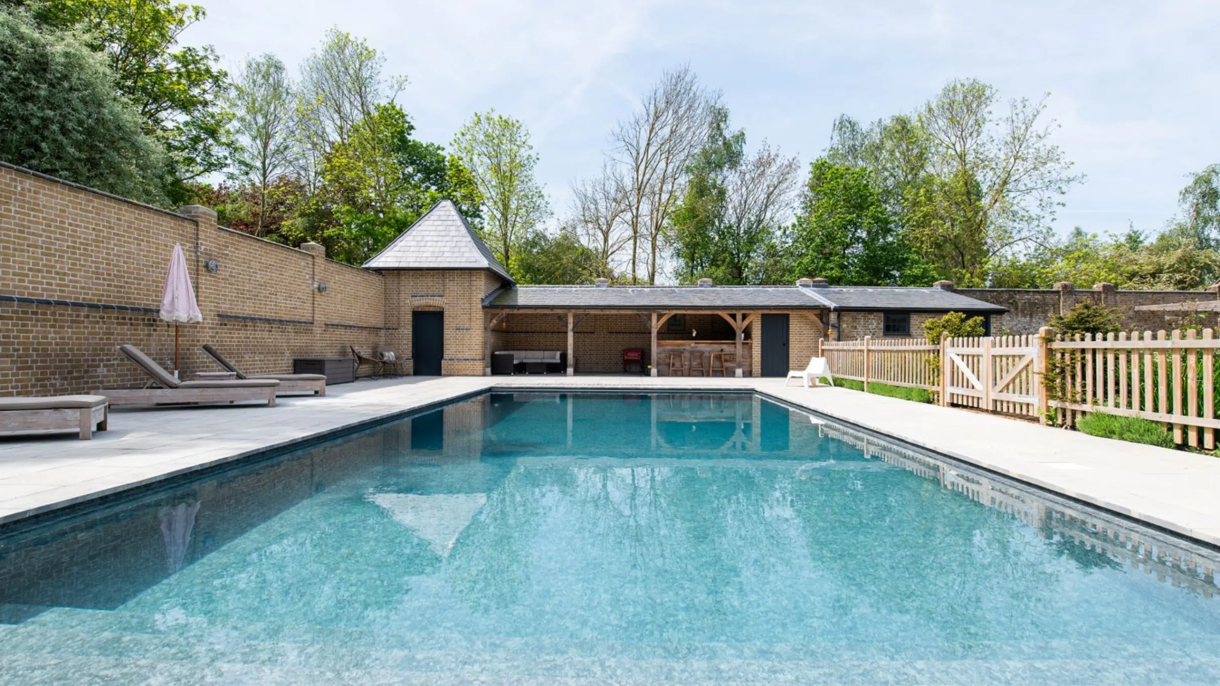 Swimming pool with sun loungers and a brick wall on one side and a picket fence on the other, with an outhouse at the back