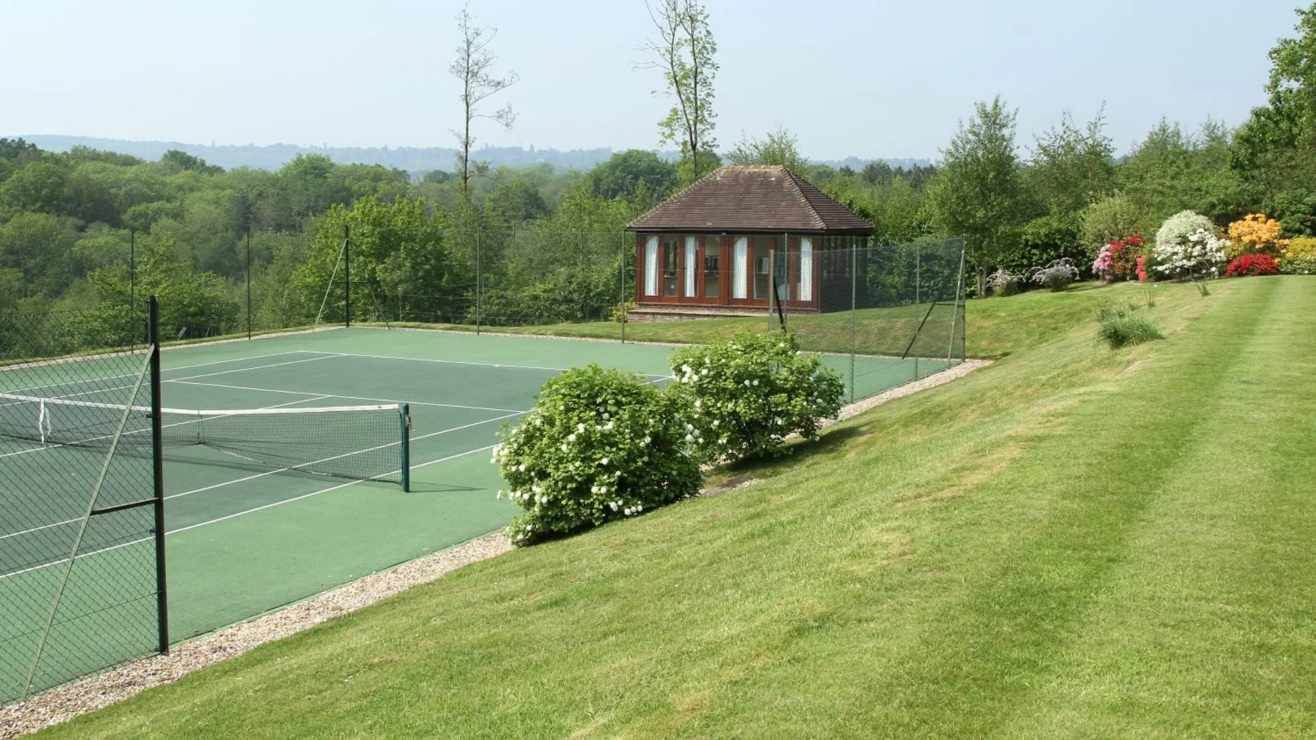 Green tennis court surrounded by green grass with trees and a brown outhouse in the background