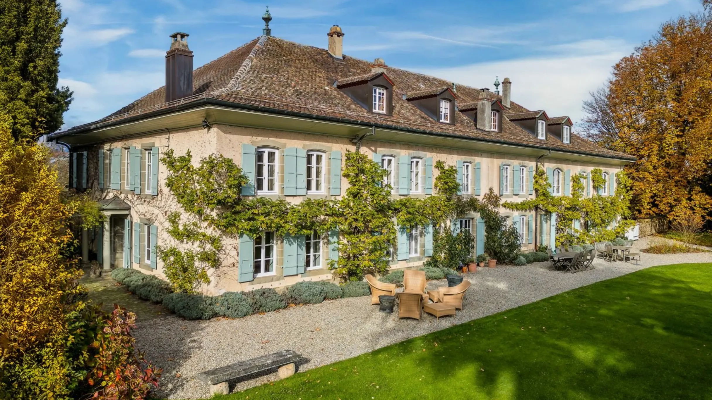 Exterior of large villa with blue shutters and front garden with table and chairs on gravel