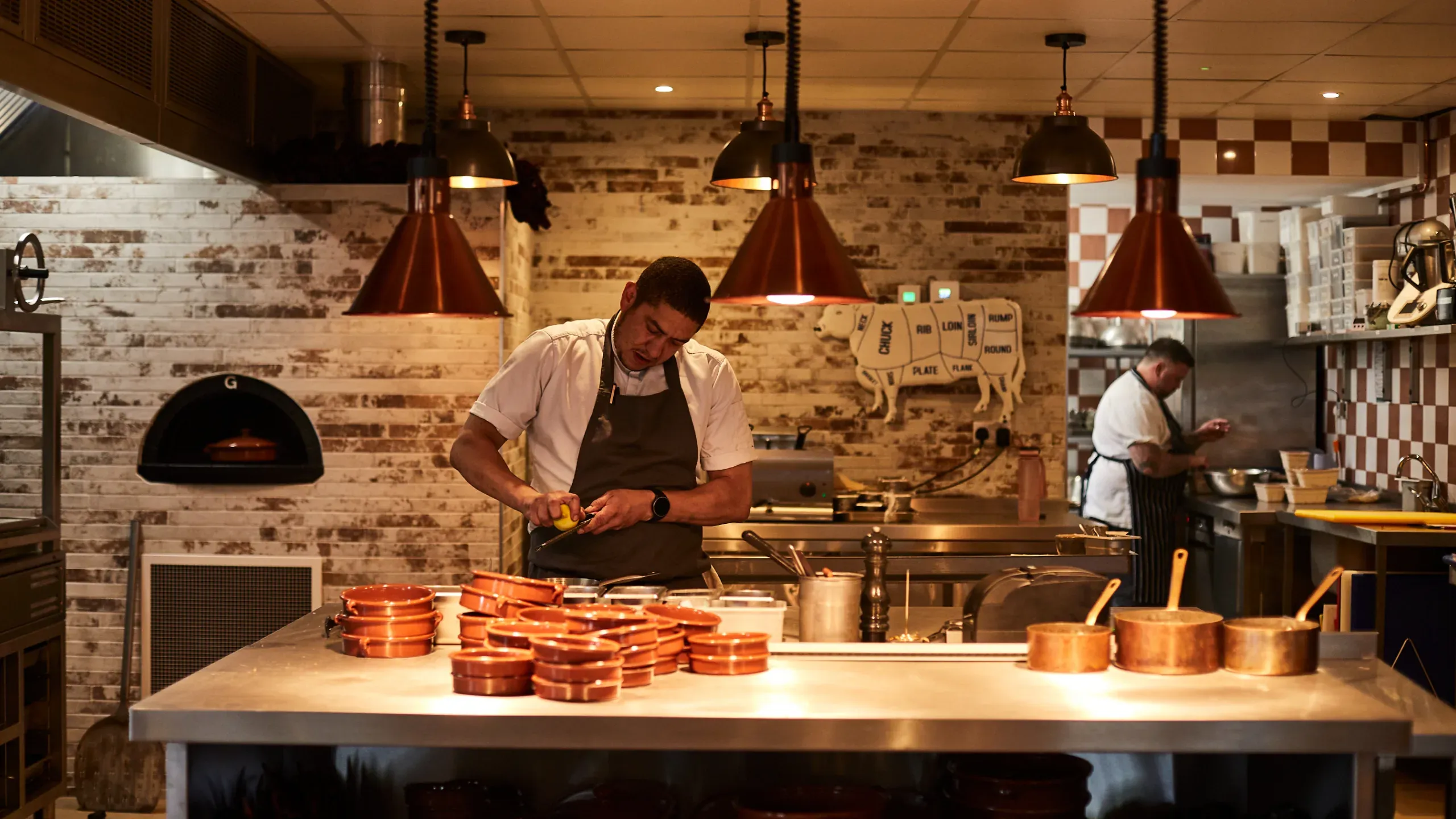 the-pot-kiln-restaurant-kitchen-with-a-man-juicing-a-lemon-surrounded-by-pots-and-pans-on-a-silver-countertop-and-another-man-cooking-in-the-background