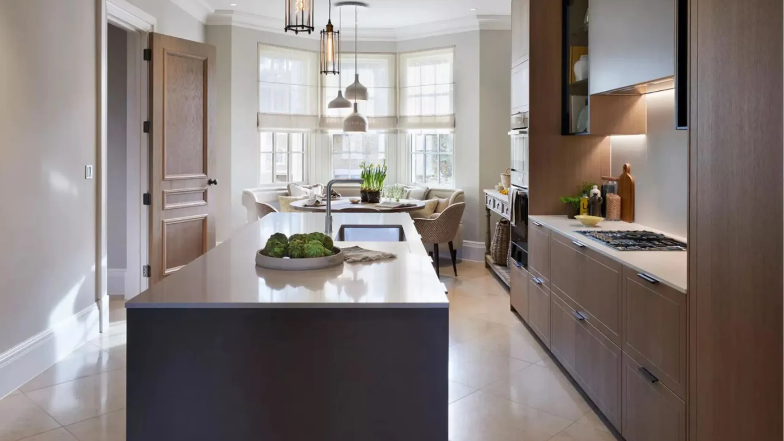 Modern kitchen with brown cabinetry a dark brown and white island, white flooring and a dining table against a window in the background