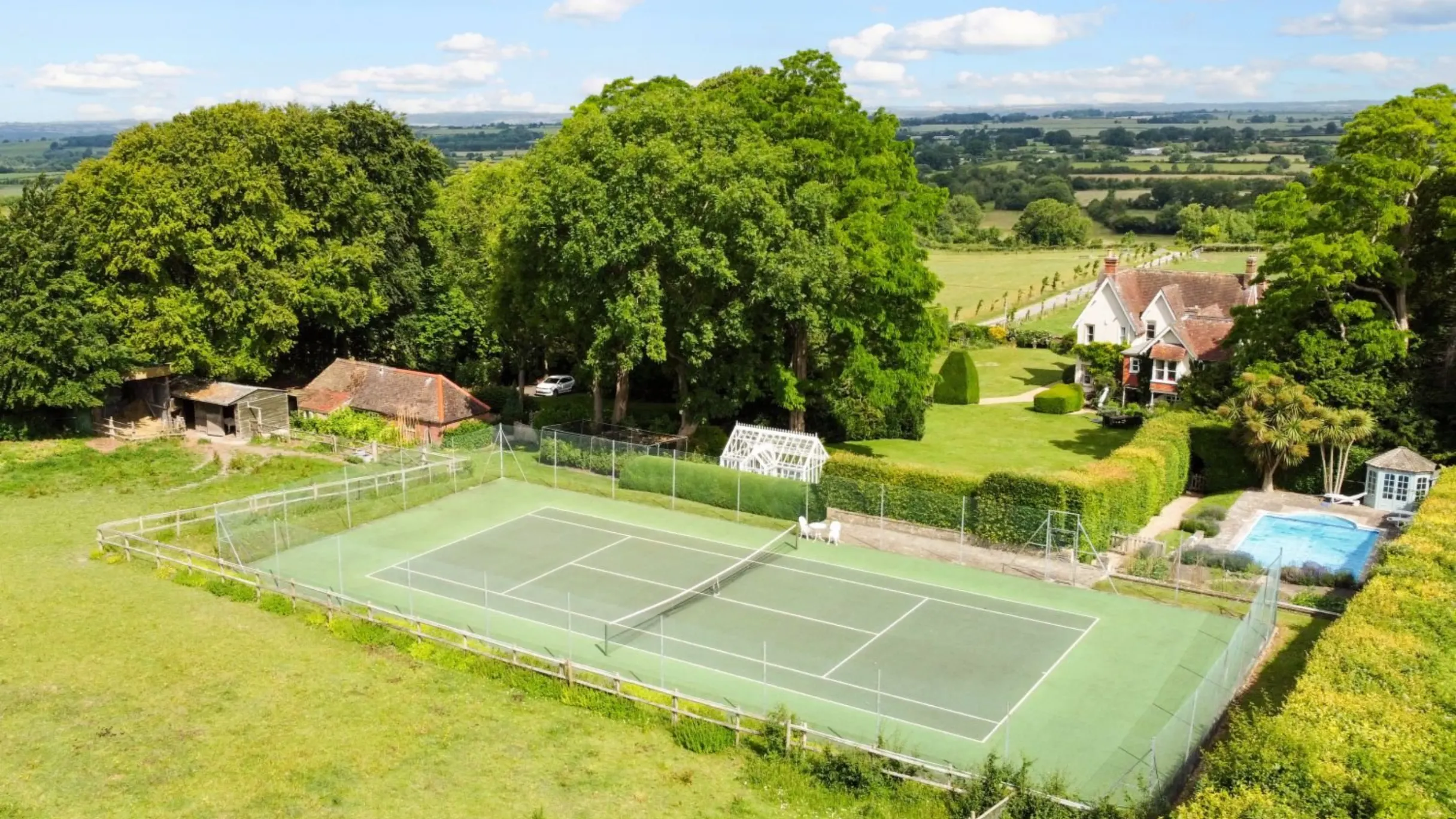 Green tennis field surrounded by green fields and trees with a swimming pool and country home in the background