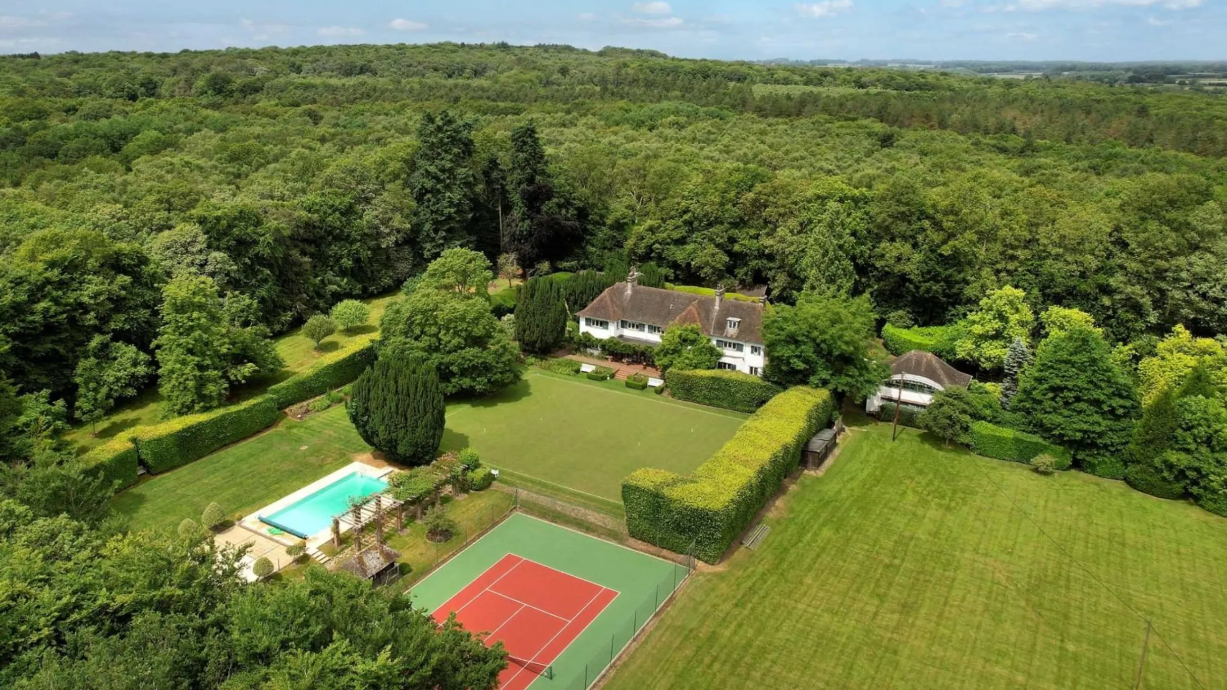 Large country home surrounded by fields and trees with an outdoor swimming pool and green and red tennis court