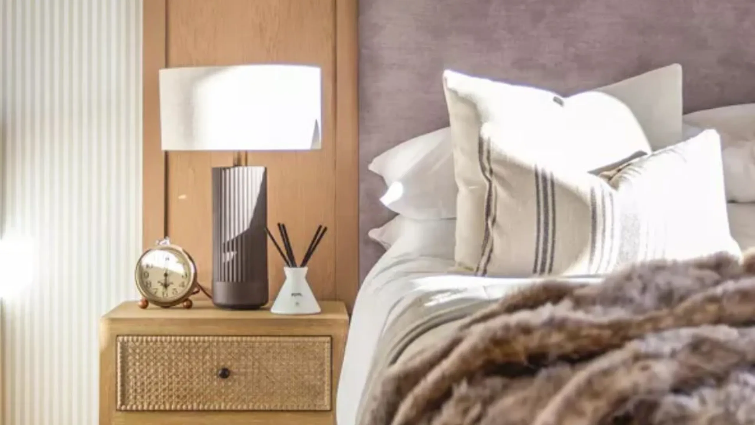 Bedroom with brown bedside table with a lamp on and bed with white sheets and lilac headboard
