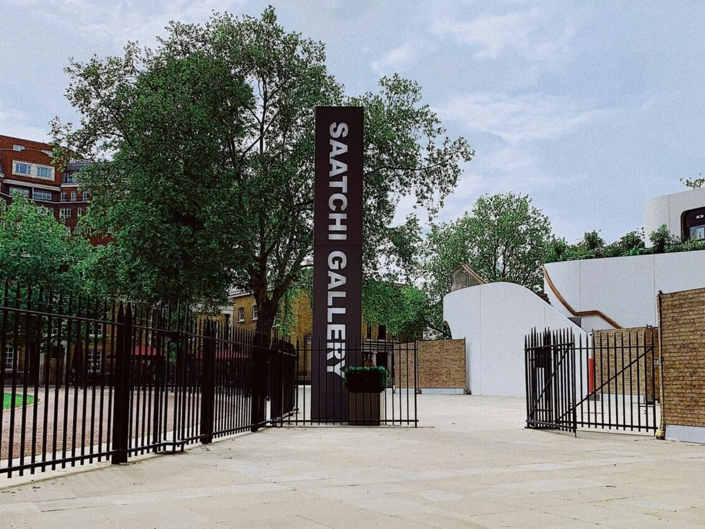 Black and white Saatchi Gallery sign in front of black metal gates with trees and buildings in the background