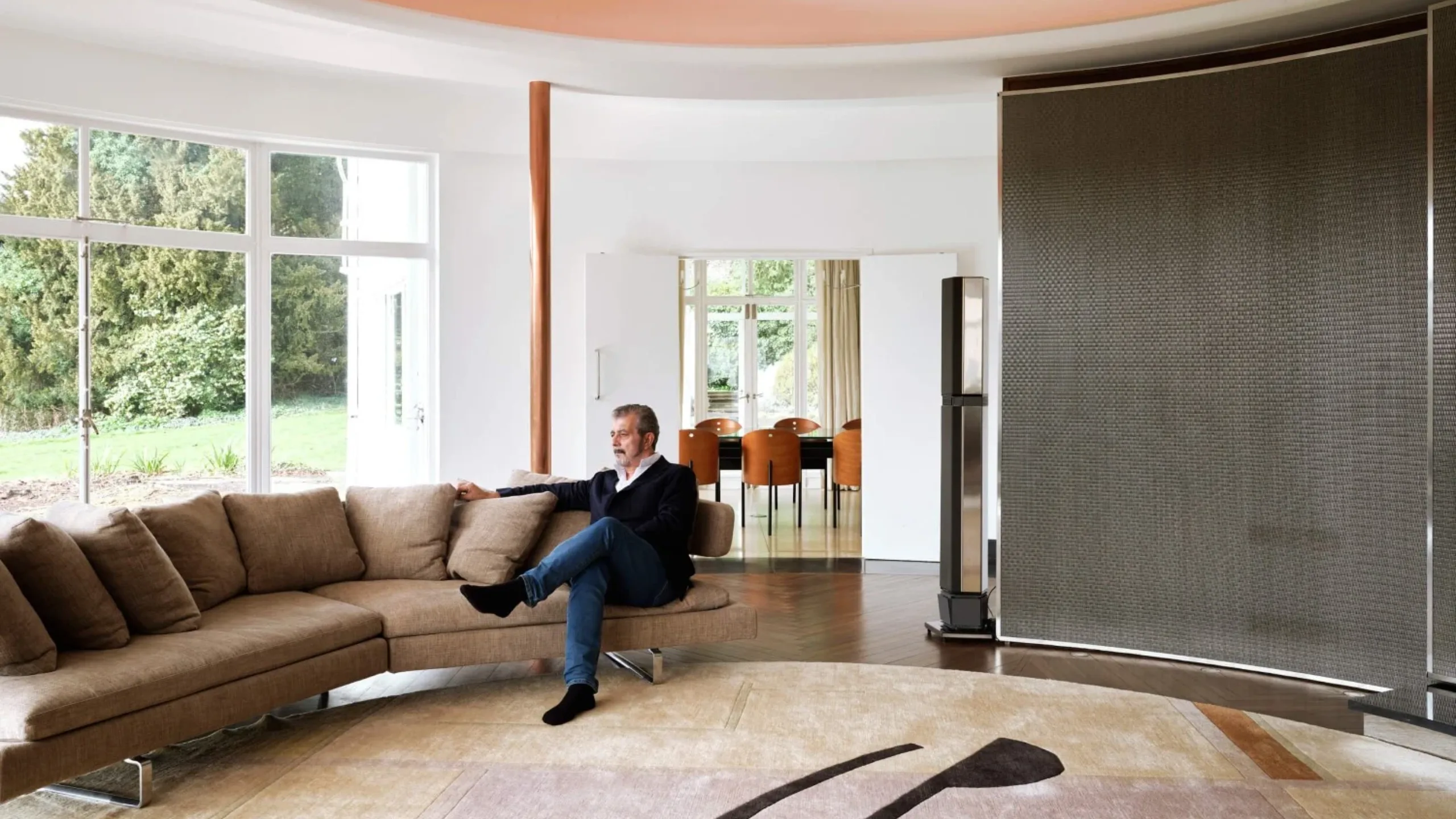 A man sitting on a beige sofa in a curved living room with white walls, beige rug and large windows
