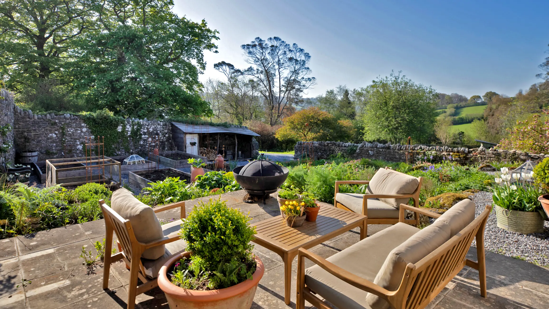 Patio with wooden table and chairs on it, surrounded by a lawn with shrubs and trees with an outhouse in the background