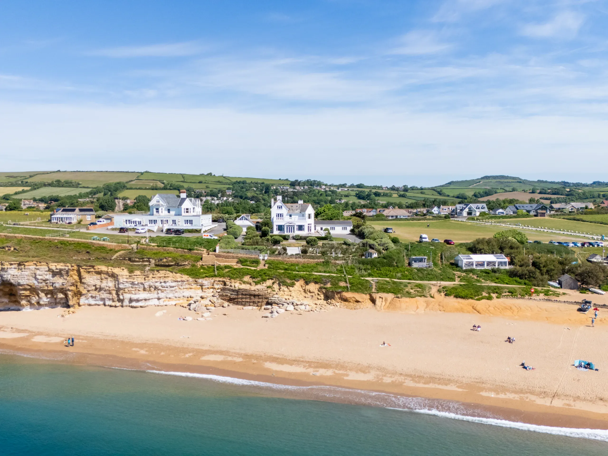 Ariel shot of coastal properties with rolling hills and trees in the background, with a beachy sand and water in the foreground