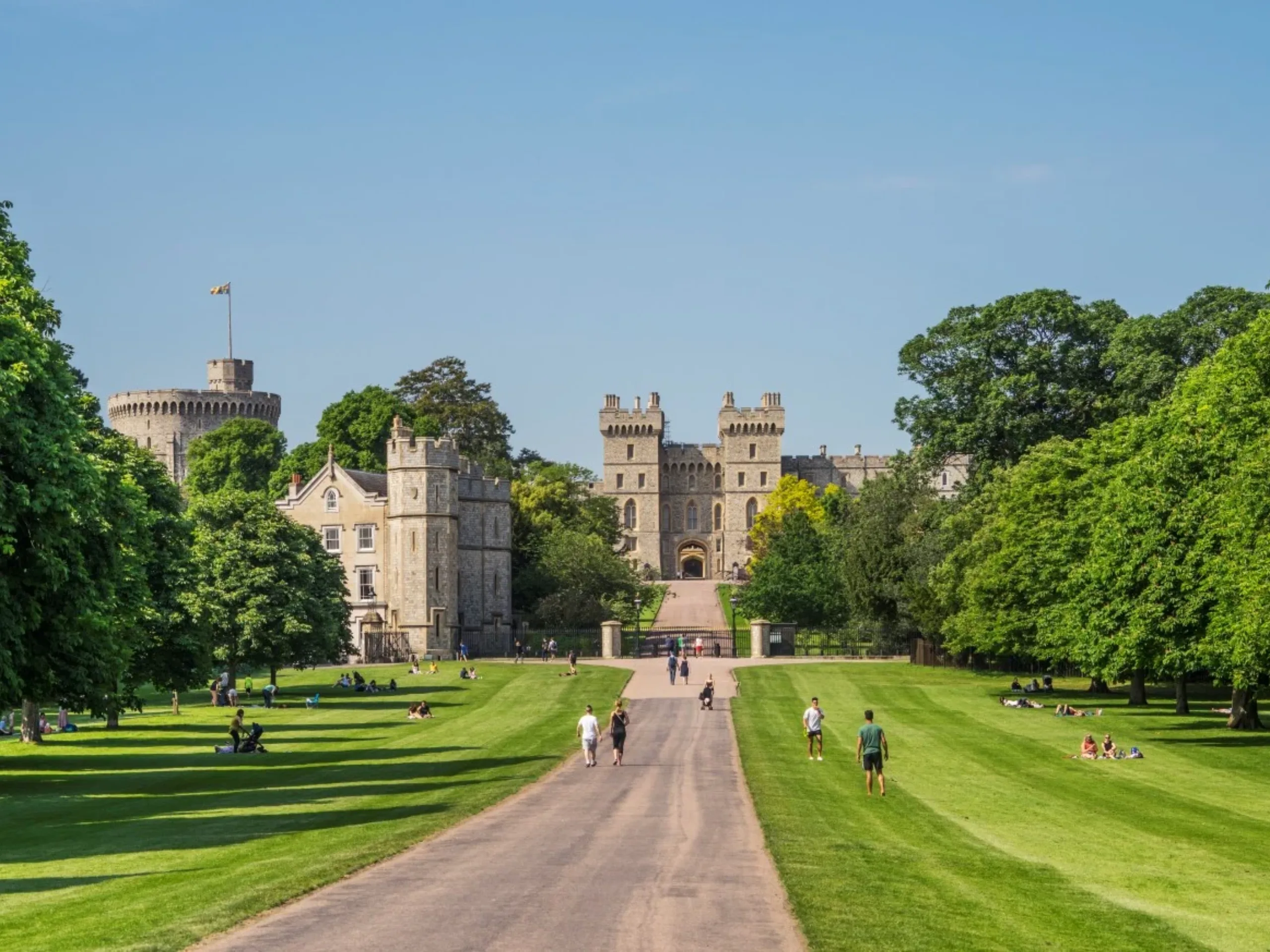 Path leading up to Windsor Castle flanked by green grass with trees in the background and people walking across the grounds