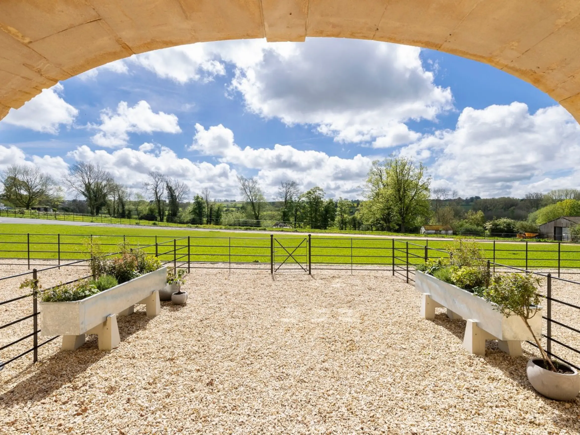 Gravel path with planters leading to grass field with trees in the distance under a blue sky