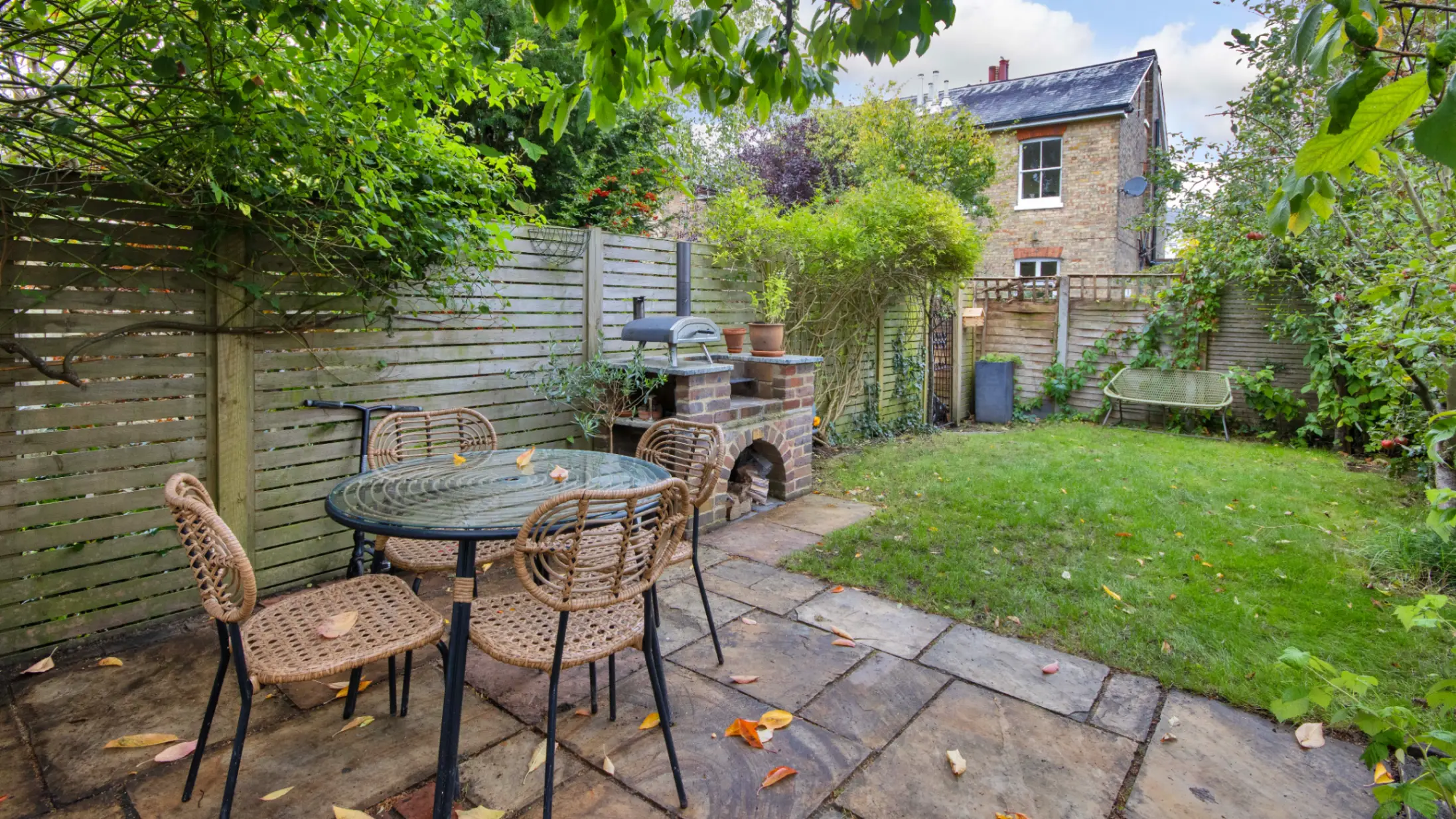 Garden with a terrace and dining table and chairs on it leading out to lawn surrounded by trees and shrubs with a small pizza oven to the side