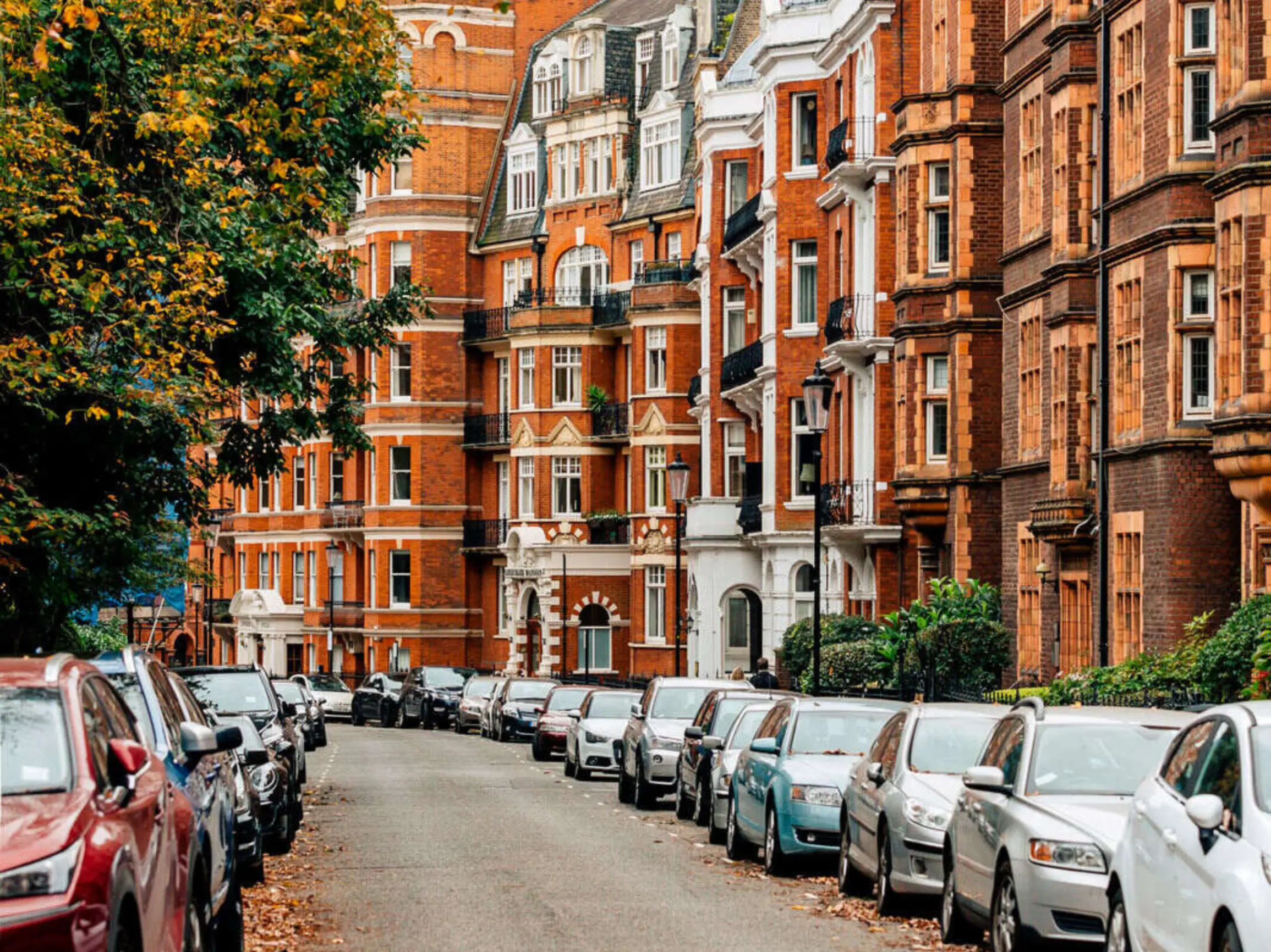 Exterior of redbrick houses on a street with trees on one side and cars on the road outside