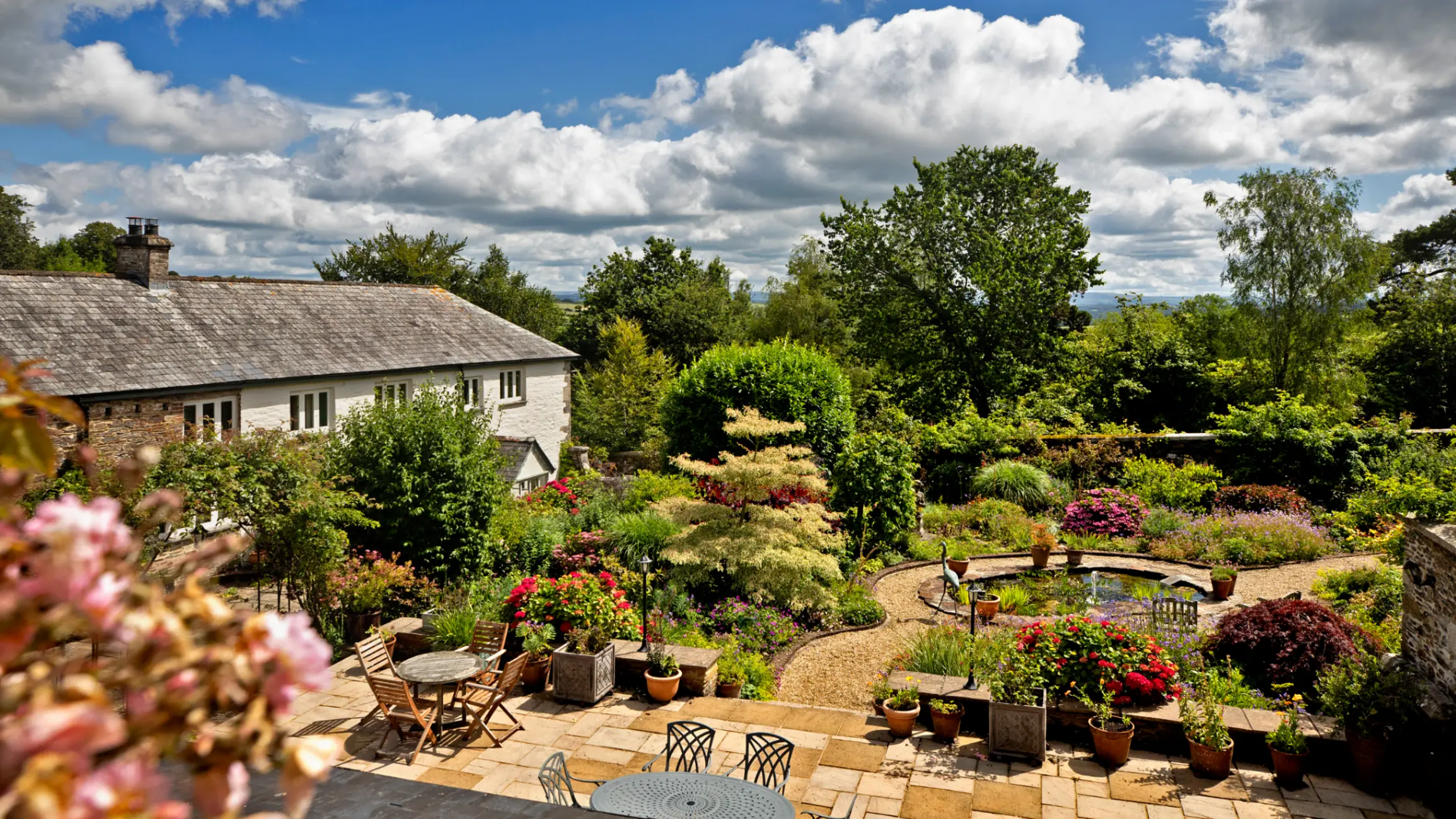 exterior of a farmhouse with a garden filled with trees, bushes, a pond and outdoor seating