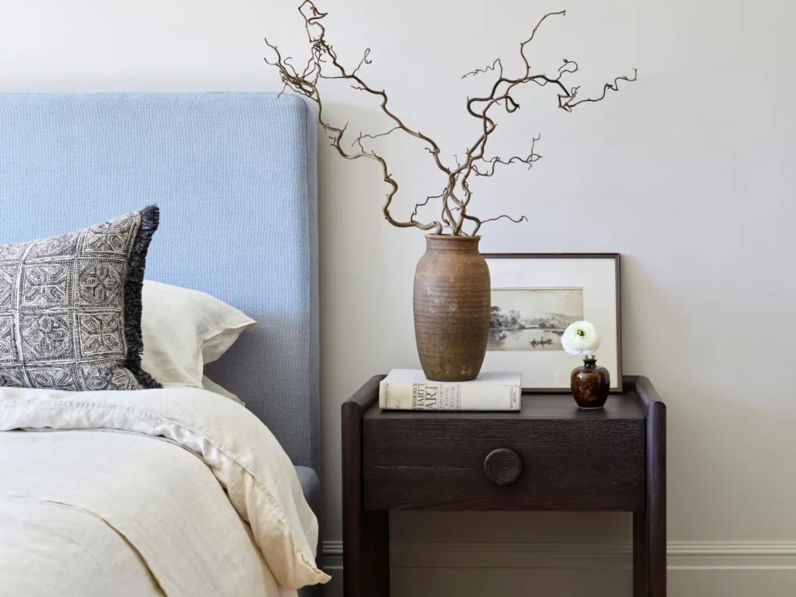 Brown bedside table with vase and books on top with the edge of a bed with a blue headboard and cream bedding next to it
