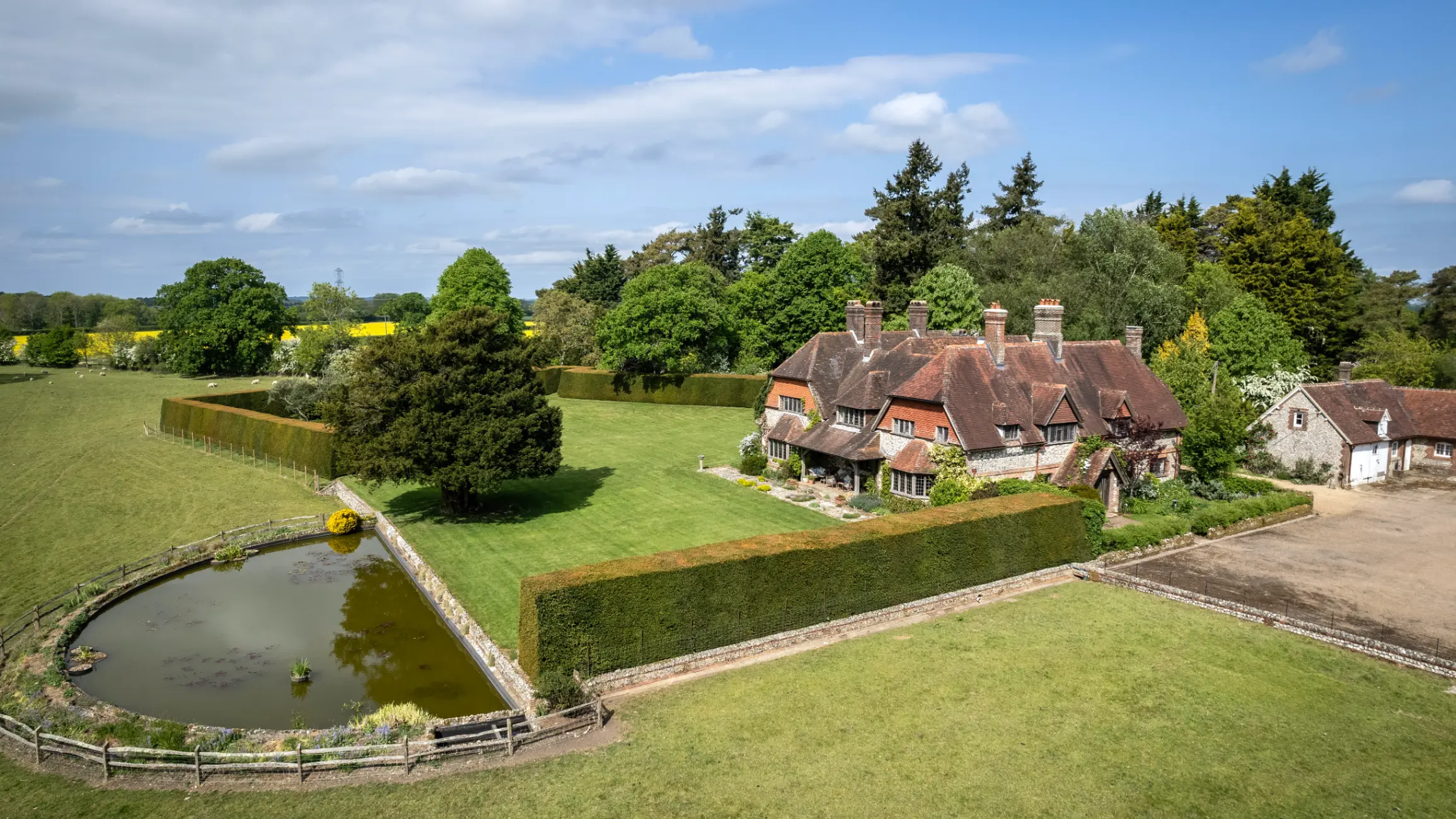 Large country home surrounded by fields and trees with a pond in front