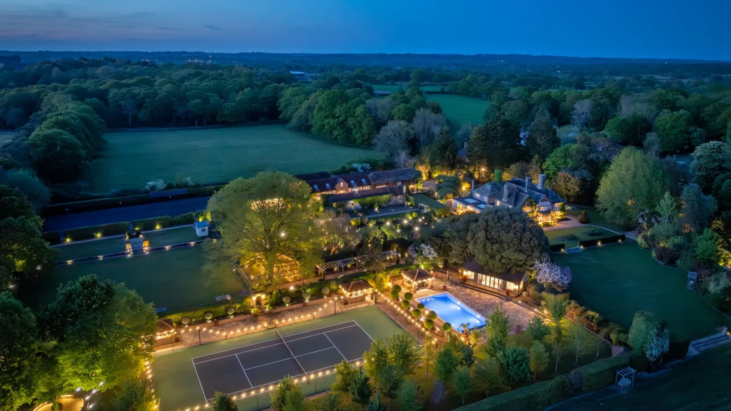 Ariel shot at night of tennis court illuminated by outdoor lights surrounded by fields and trees with a swimming pool and country house in the background