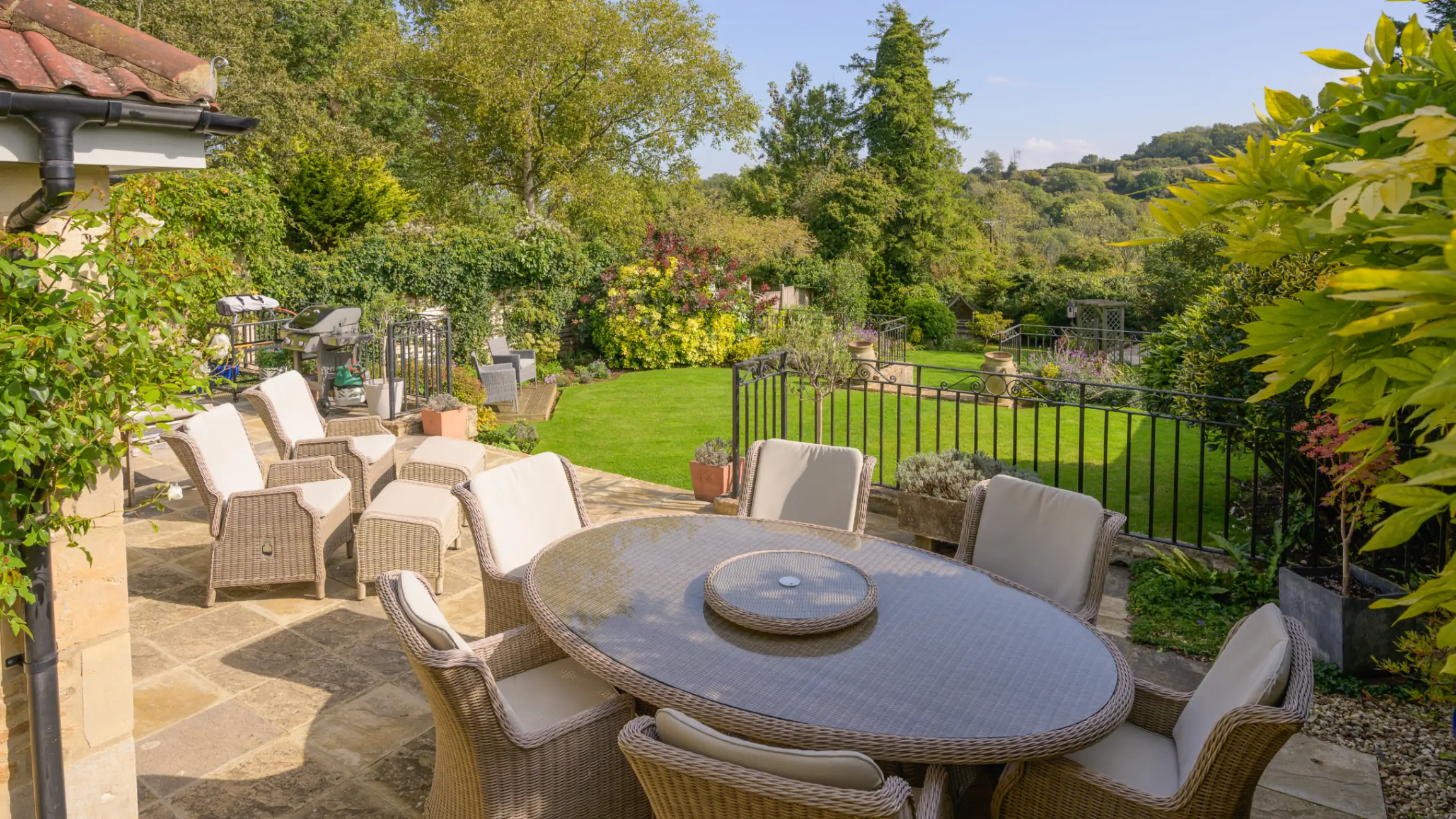 Garden with raised patio with a dining table and padded chairs and two further padded chairs looking out a lawn surrounded by shrubs and trees
