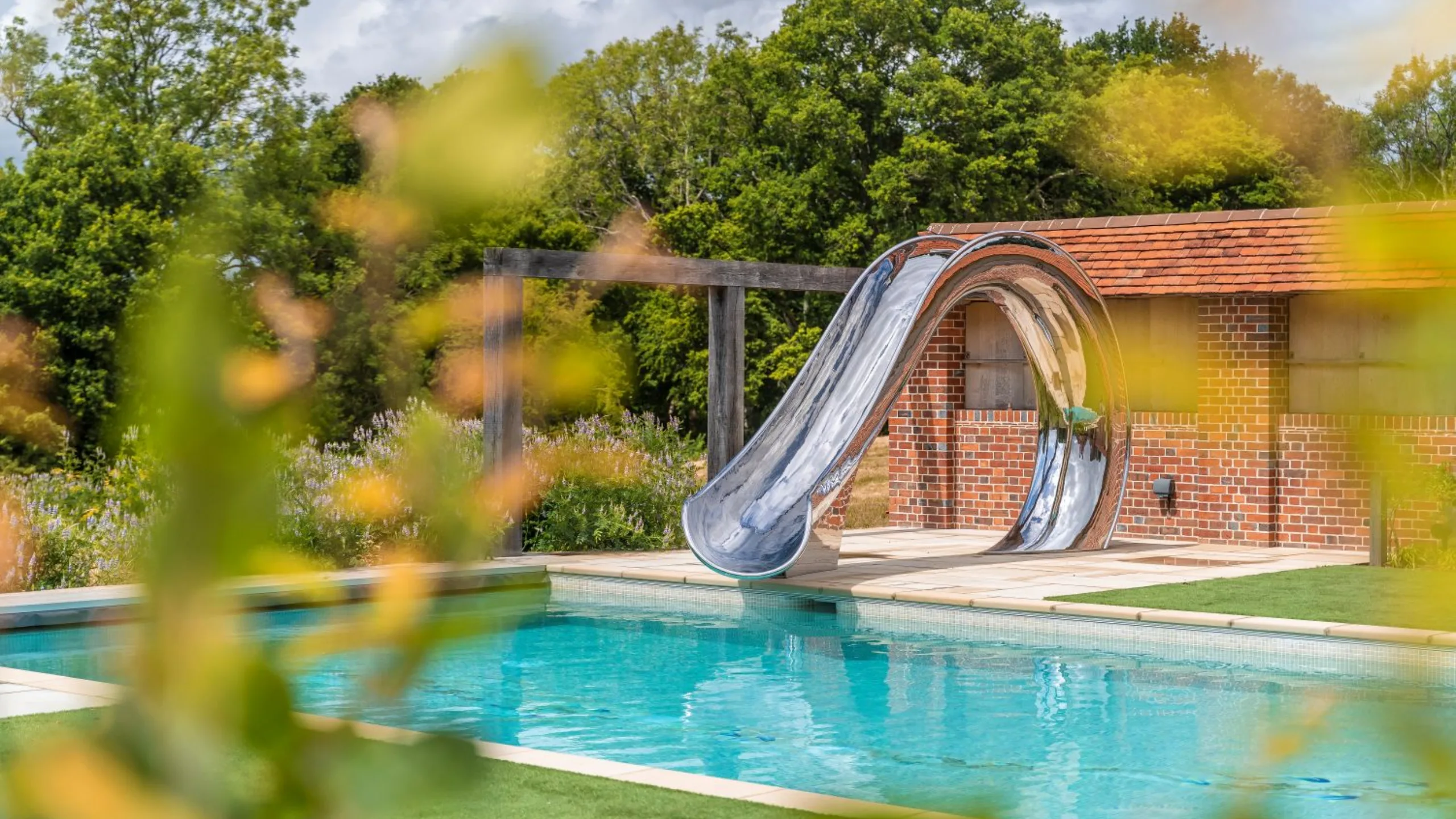 Curved silver slide going into a pool with a red brick building in the background surrounded by trees