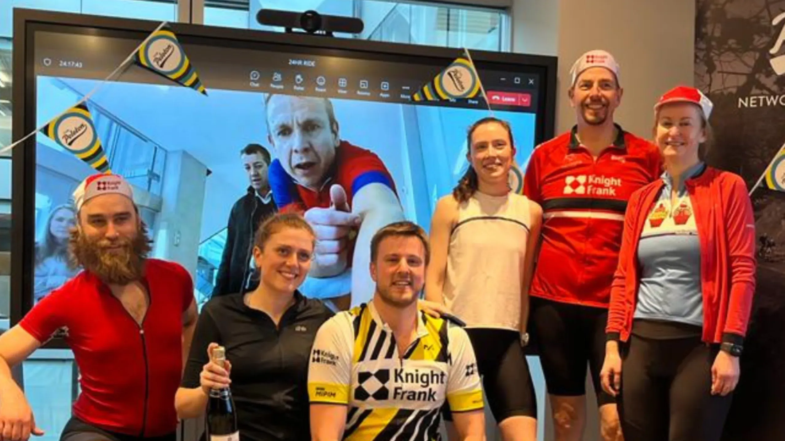 group-of-people-in-branded-knight-frank-cycling-gear-smiling-at-the-camera-in-front-of-a-large-projector-screen-with-a-man-looking-out