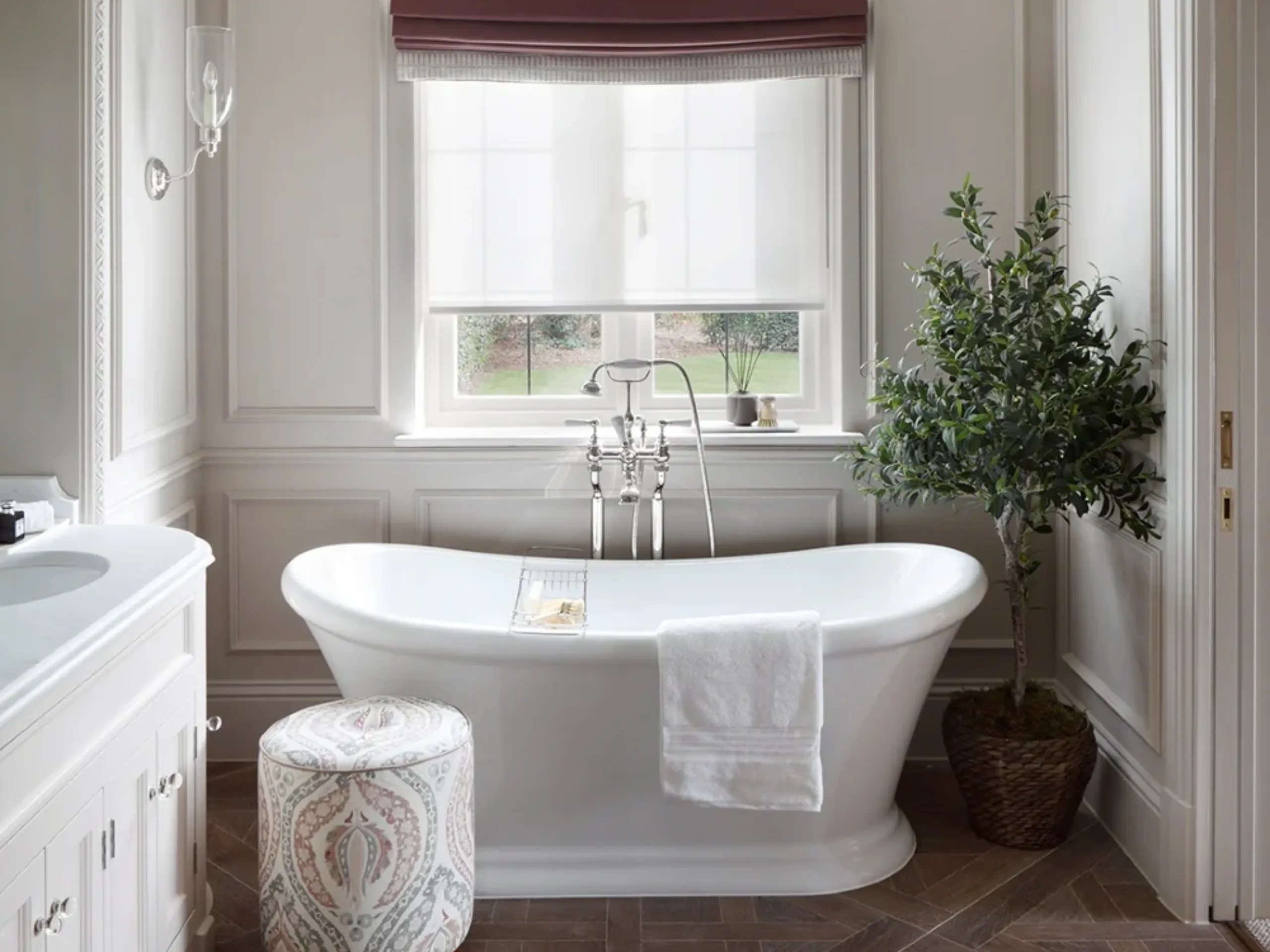 Bathroom with freestanding bath, with a window above, white walls, patterned stool and potted plant