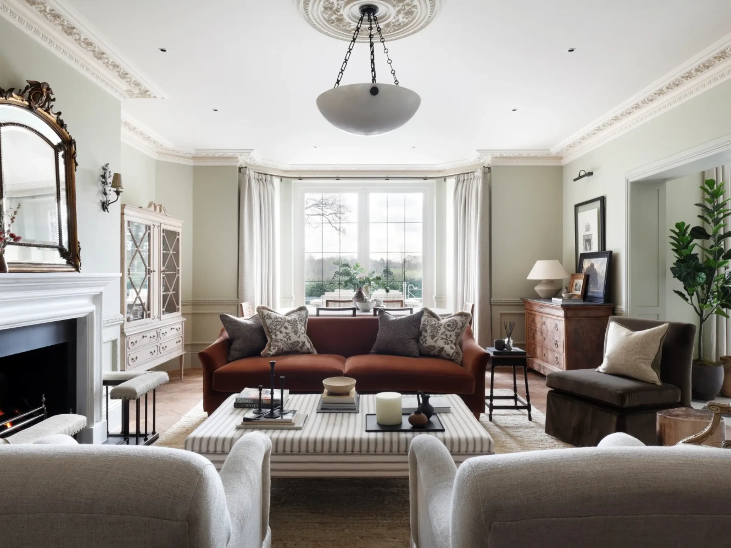 period living room with cream armchairs, a red sofa, large ottoman, fireplace and large bay windows 