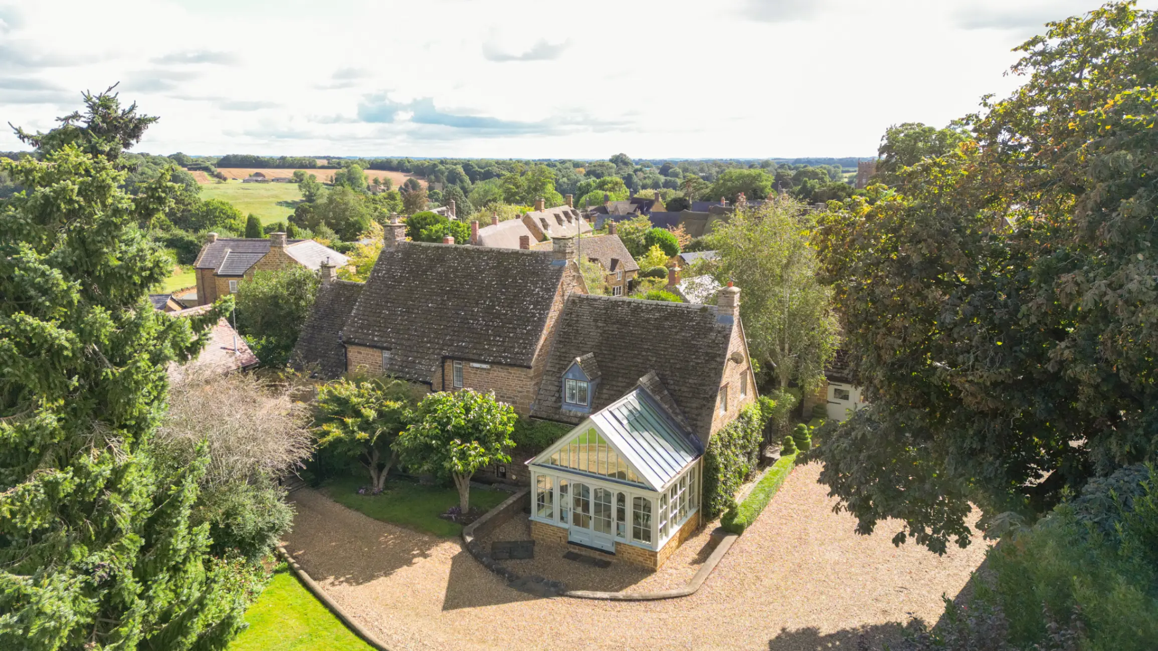 Exterior of a country house with a glass conservatory, surrounded by green fields and mature trees