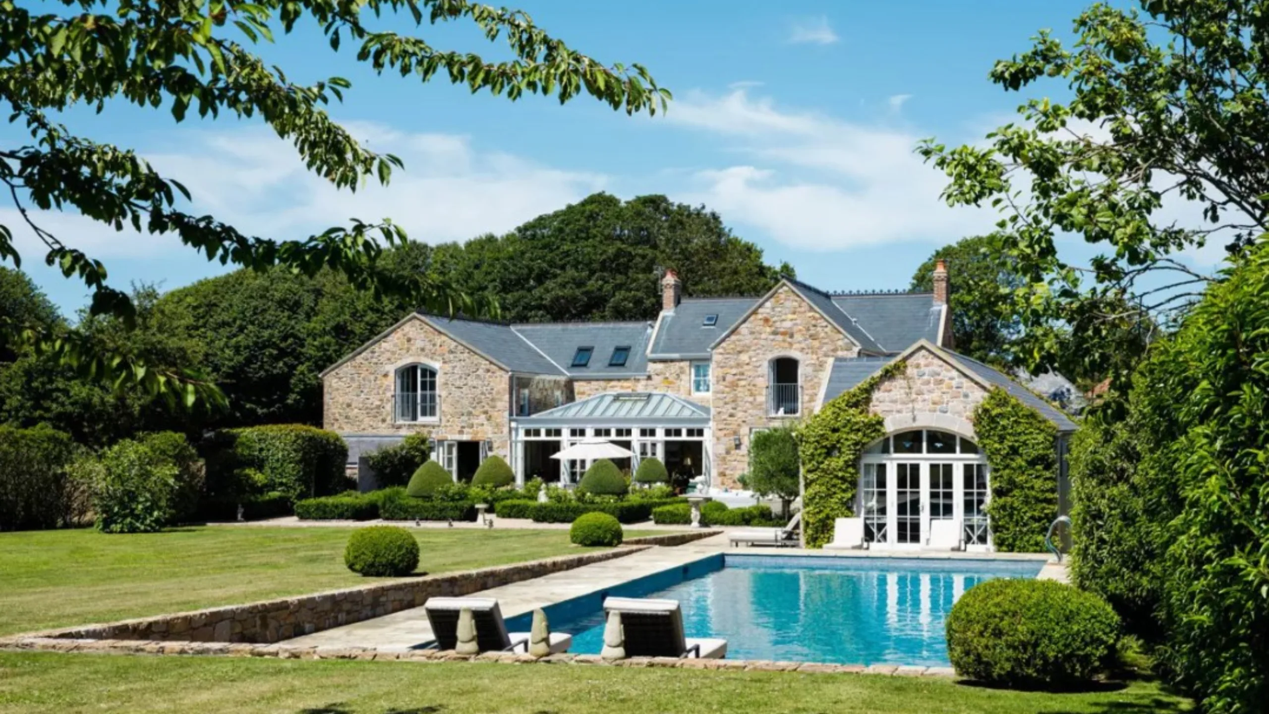 Exterior of a brick country house with a green garden, mature trees and a swimming pool in the foreground