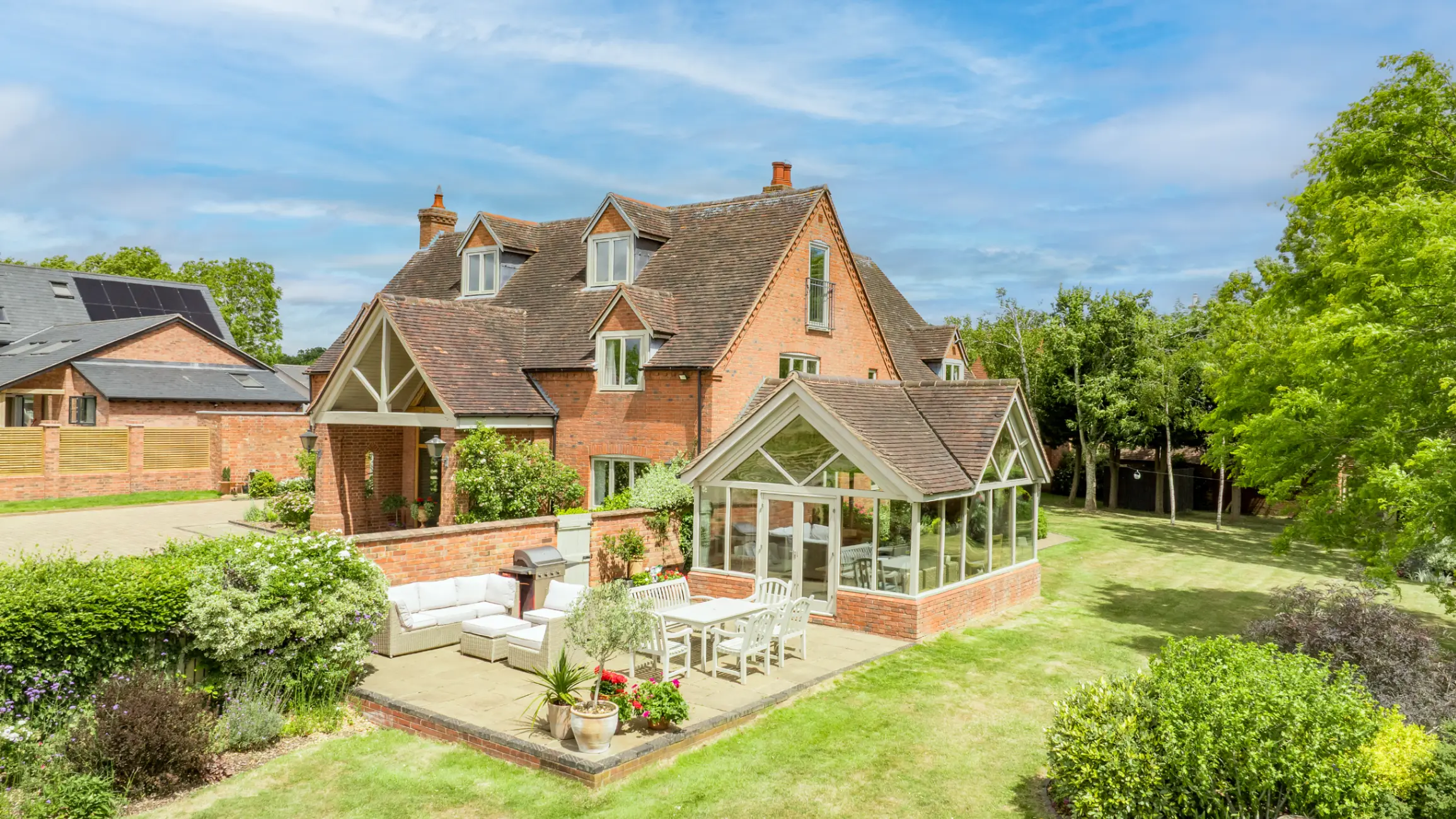 Exterior of a large red brick house with a patio and glass conservatory, surrounded by a garden with mature trees and bushes