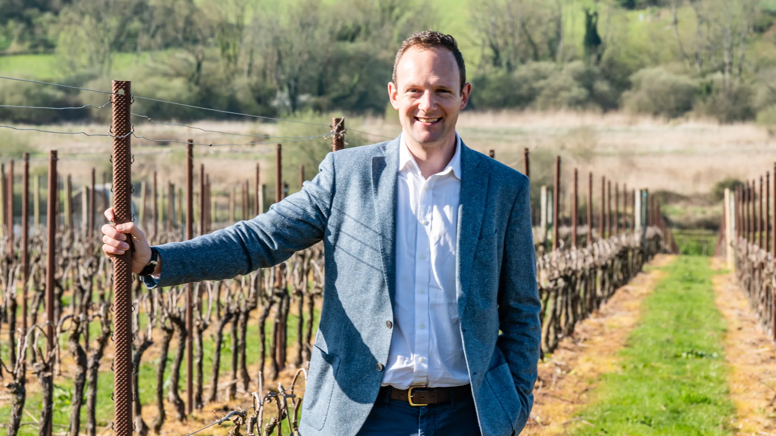 man-in-a-suit-holding-a-pole-in-a-vineyard-with-hills-in-the-background