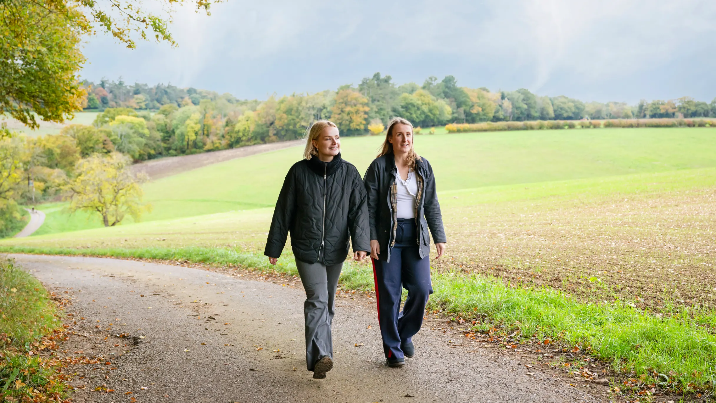 two-women-walking-on-a-path-with-green-fields-and-trees-in-the-background