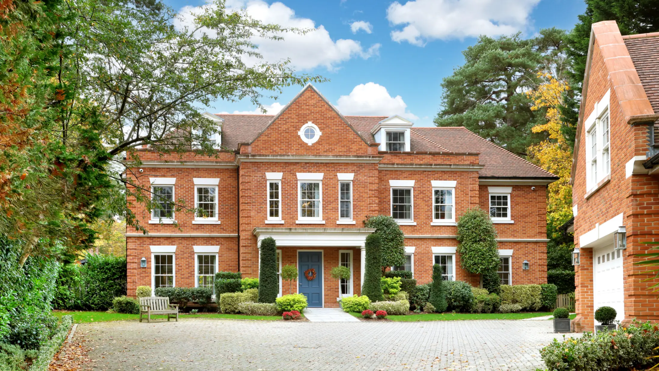 Exterior of a large red brick house with a large paved drive and flanked by bushes and trees