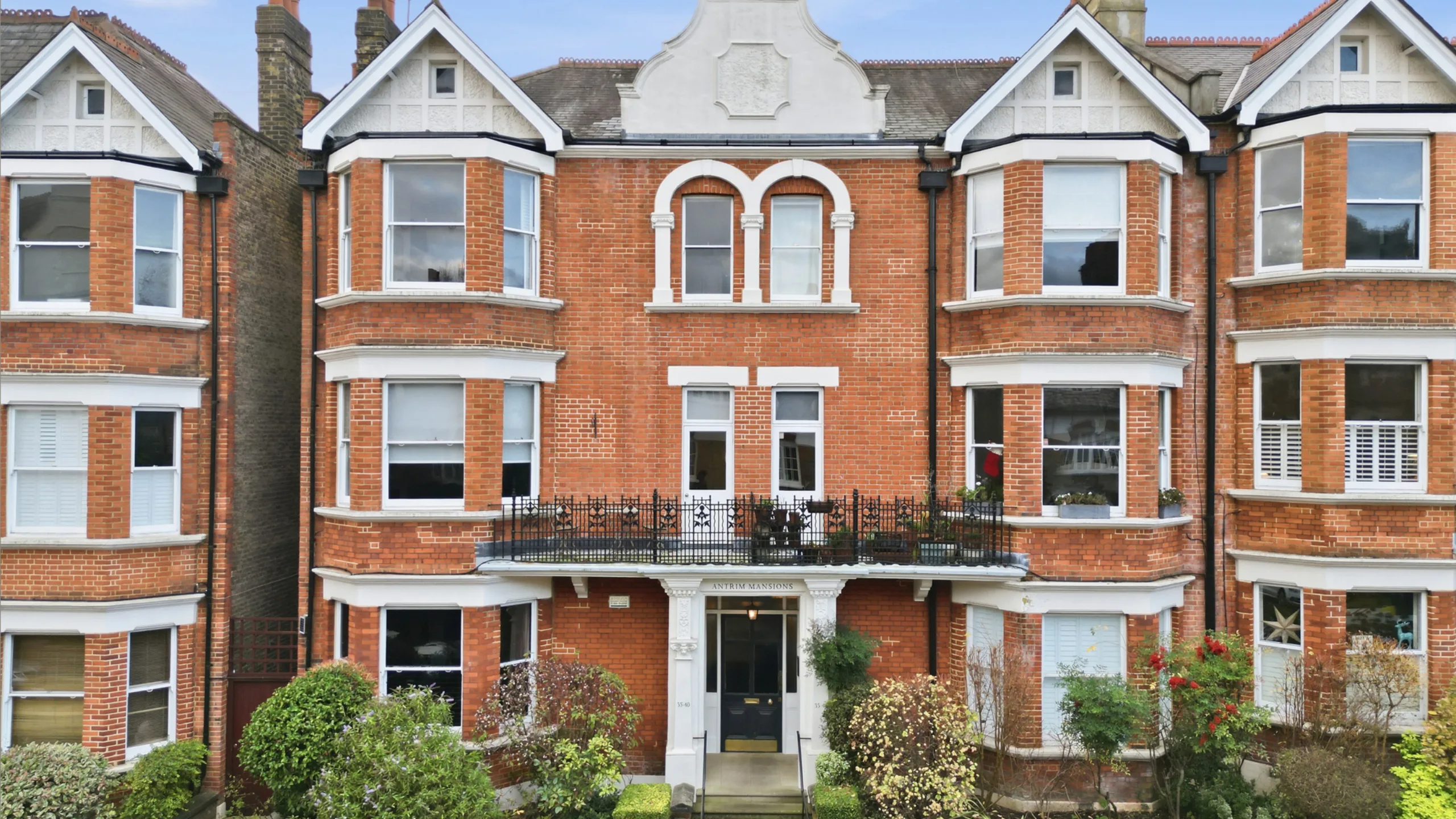 Exterior of a red brick apartment block with a black balcony and bushes in front