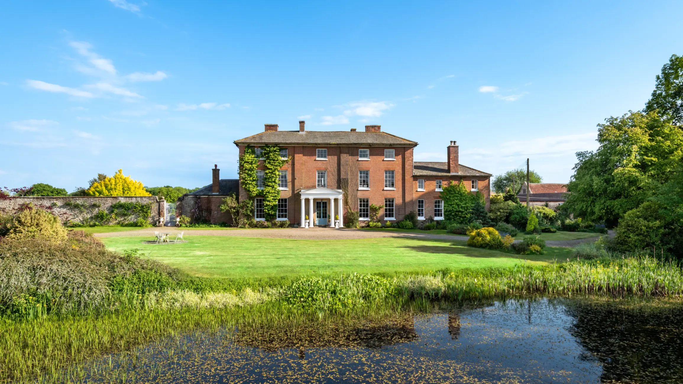 Exterior of large country house, surrounded by mature trees and a large lawn with a pond at the front