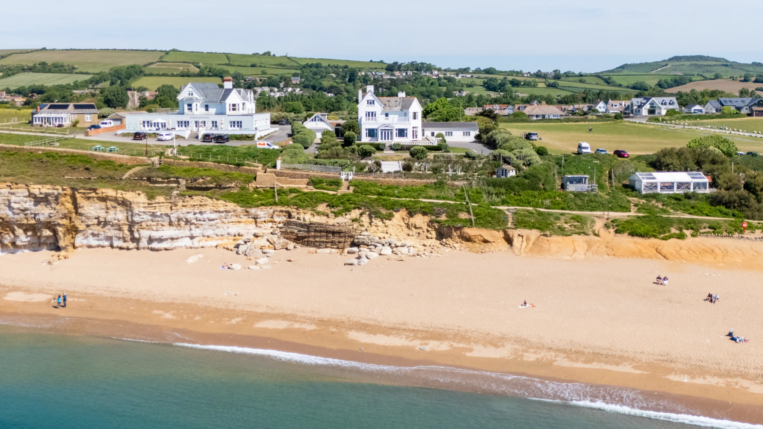 Aerial shot of large white house on a cliff amongst multiple green fields with other houses in the background and a beach in the foreground 