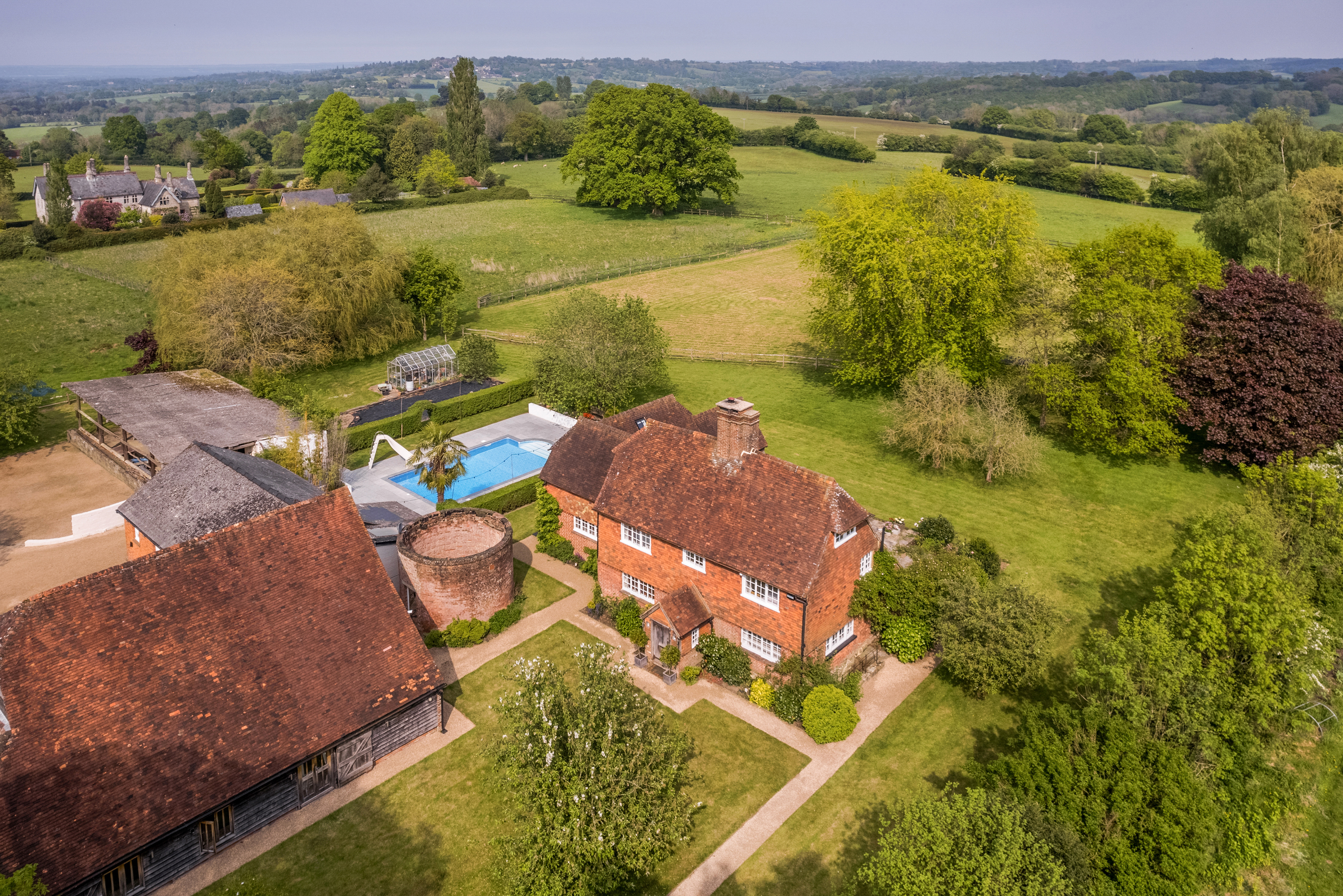 Exterior of farmhouse, surrounded by a swimming pool, outhouses and fields with mature trees