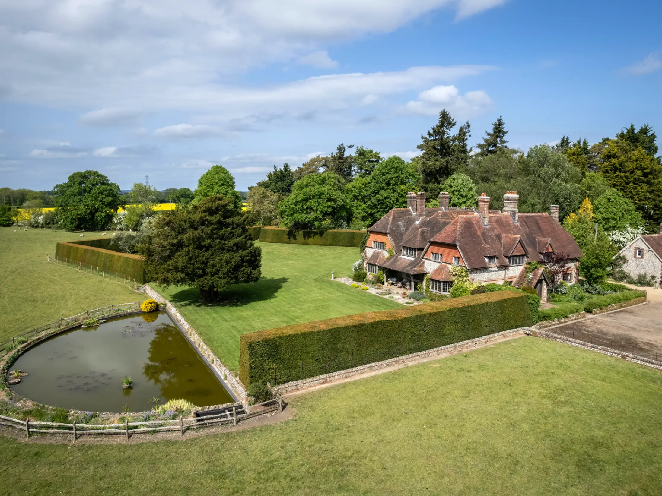 Exterior of large farmhouse surrounded by a small pond, mature trees and green fields