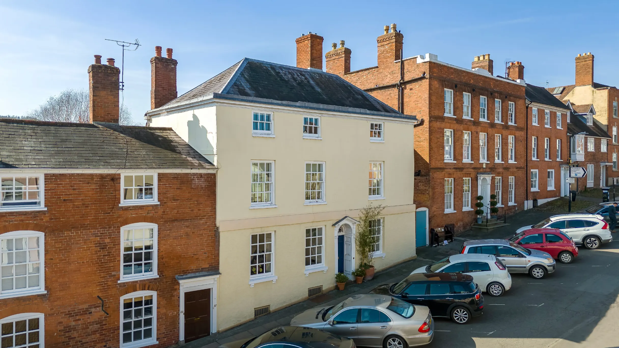 Exterior of a row of houses with a cream house in the middle flanked by red brick houses, and cars parked in front