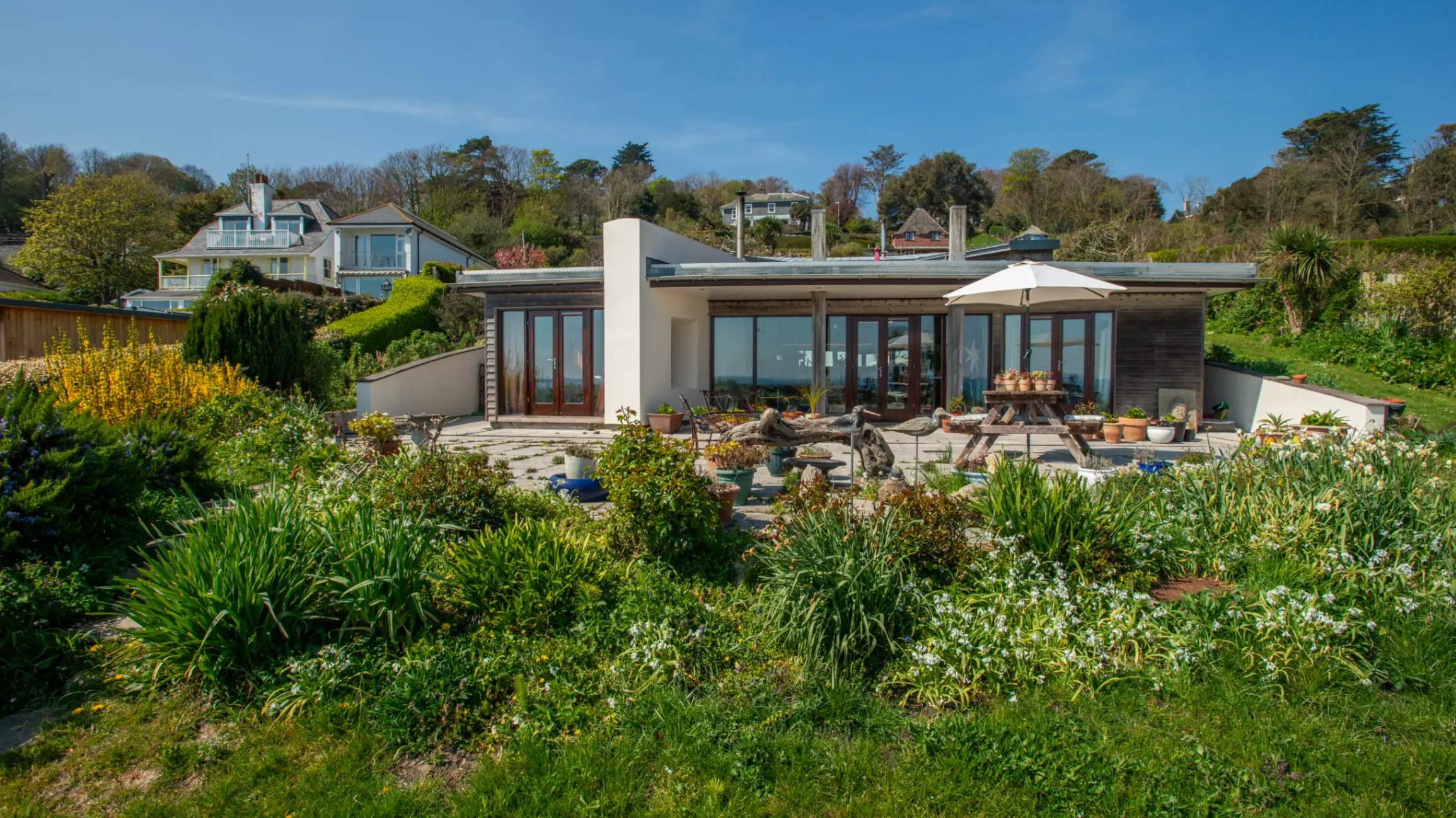 Exterior of a modern home with large glass windows, surrounded by a garden with a patio and green grass and bushes 
