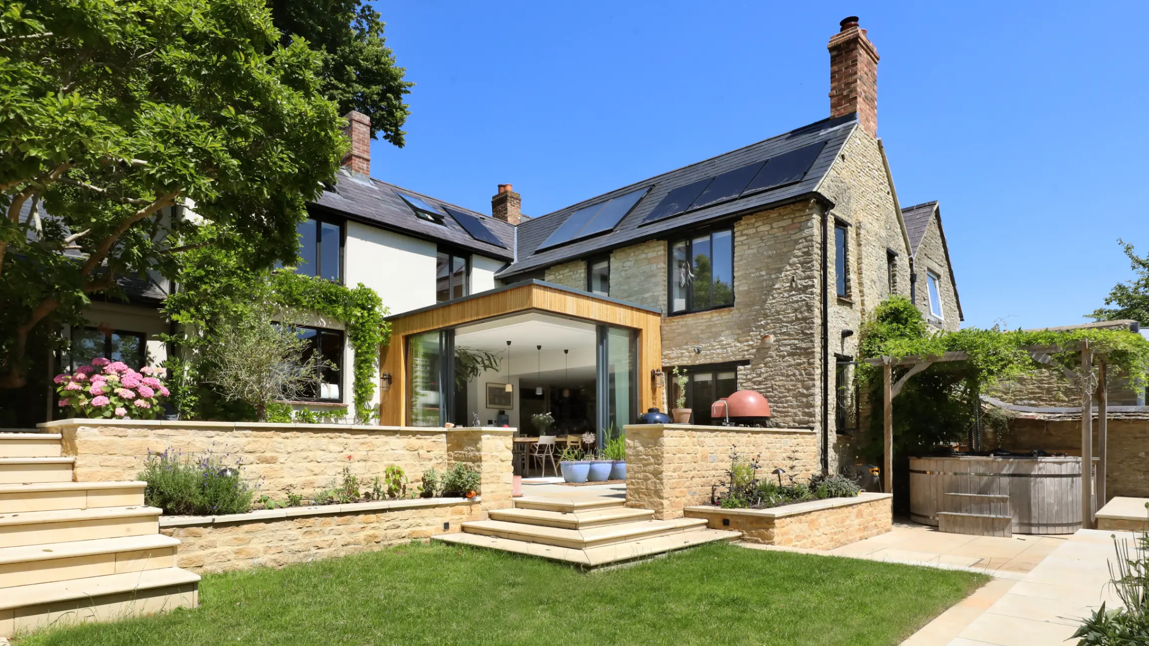 Exterior of modernised period home with glass extension, cream-bricked main house, and cream garden wall and steps leading to a garden with a hot tub