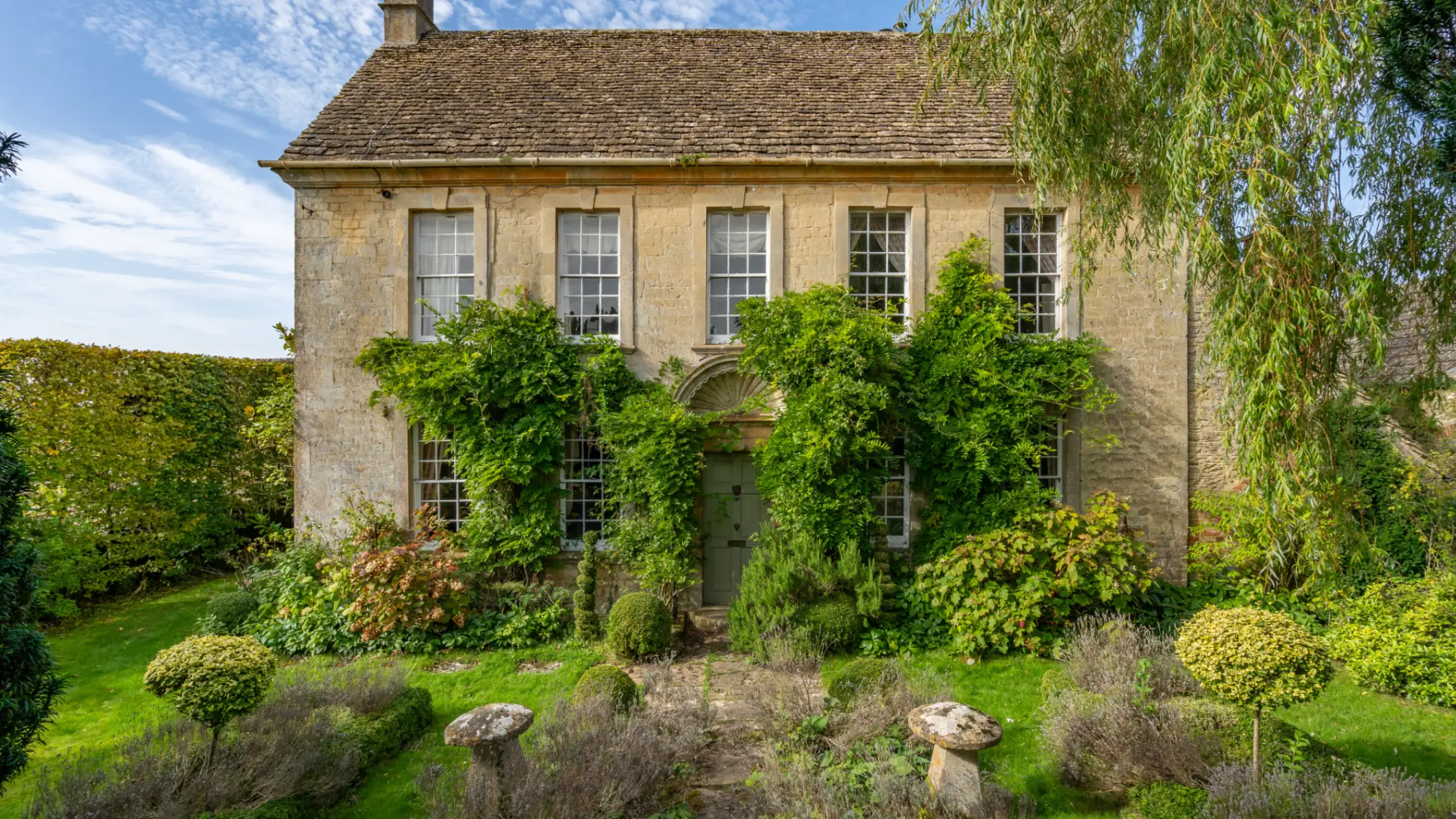 Exterior of a Georgian country house made from beige Cotswold stone with foliage growing up the walls and a front garden with bushes