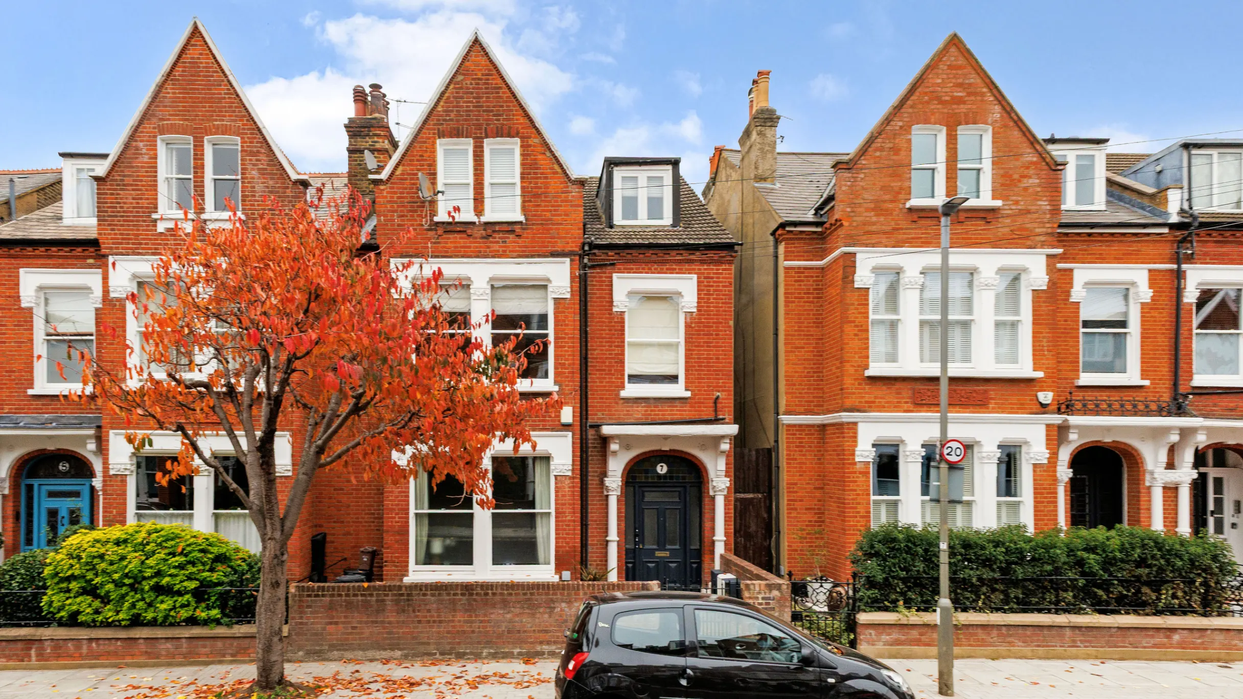 A row of a few red brick terraced houses with a black car and a red leaf tree in front 