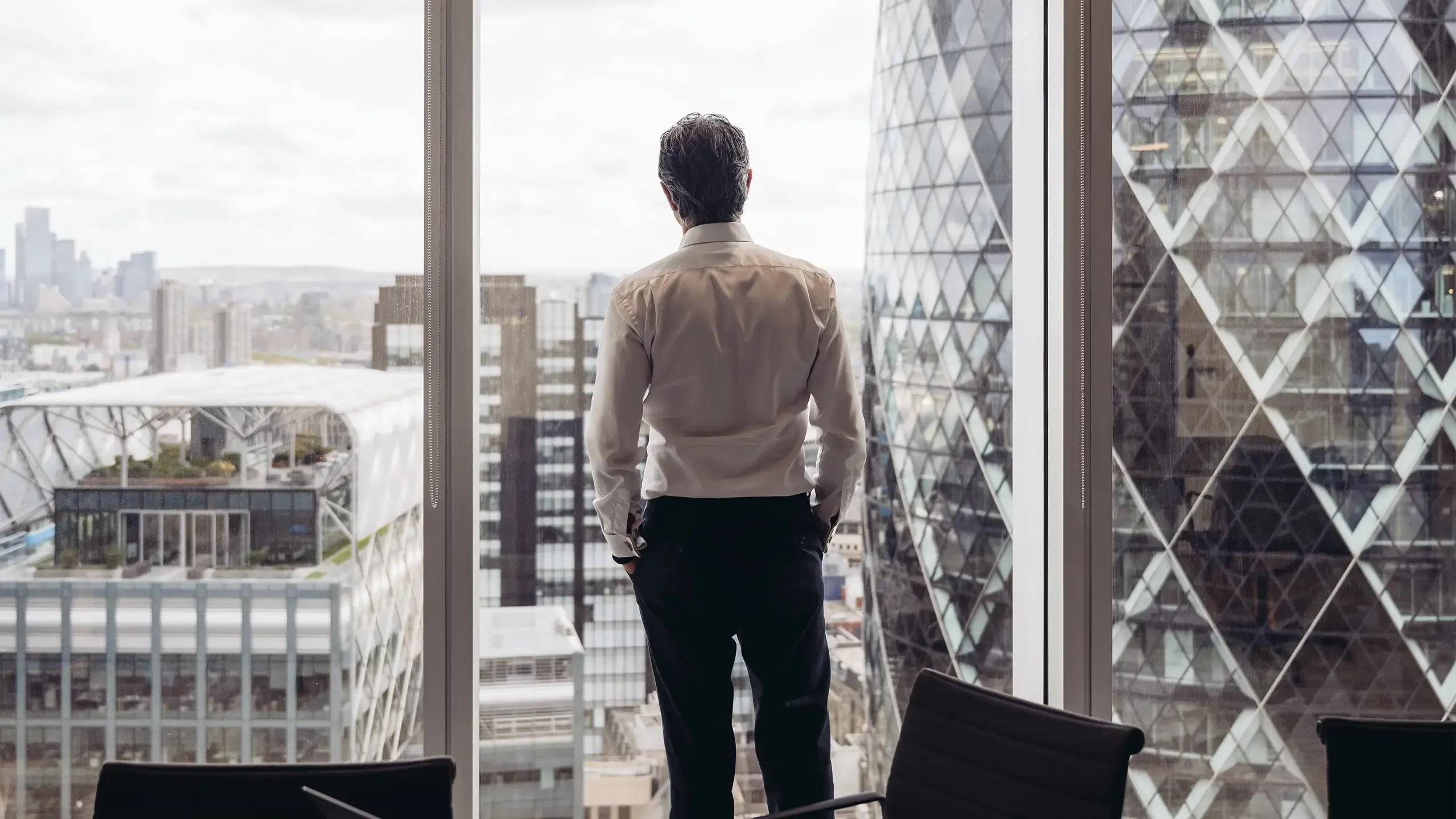 Rear view across conference table in modern board room of executive in early 40s standing with hands in pockets and looking at London’s financial district.