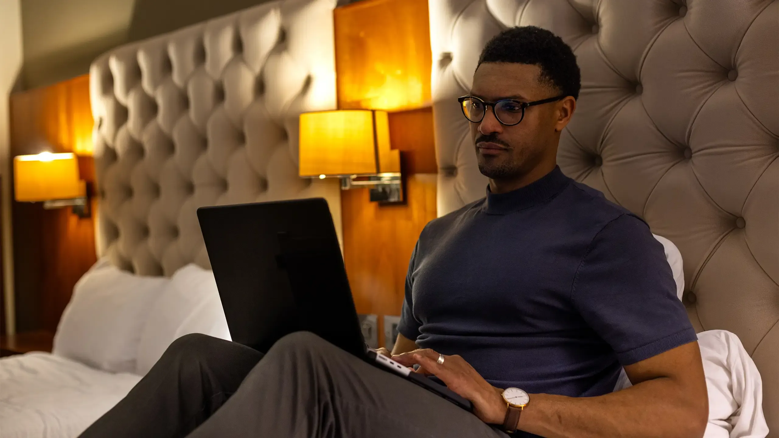 A shot of a mid-adult male sitting on a bed at nighttime in a hotel room in Newcastle Upon Tyne, North East England. He has a laptop in front of him as he works remotely by lamplight whilst away from home. He has a serious expression and is wearing glasses and casual clothing.