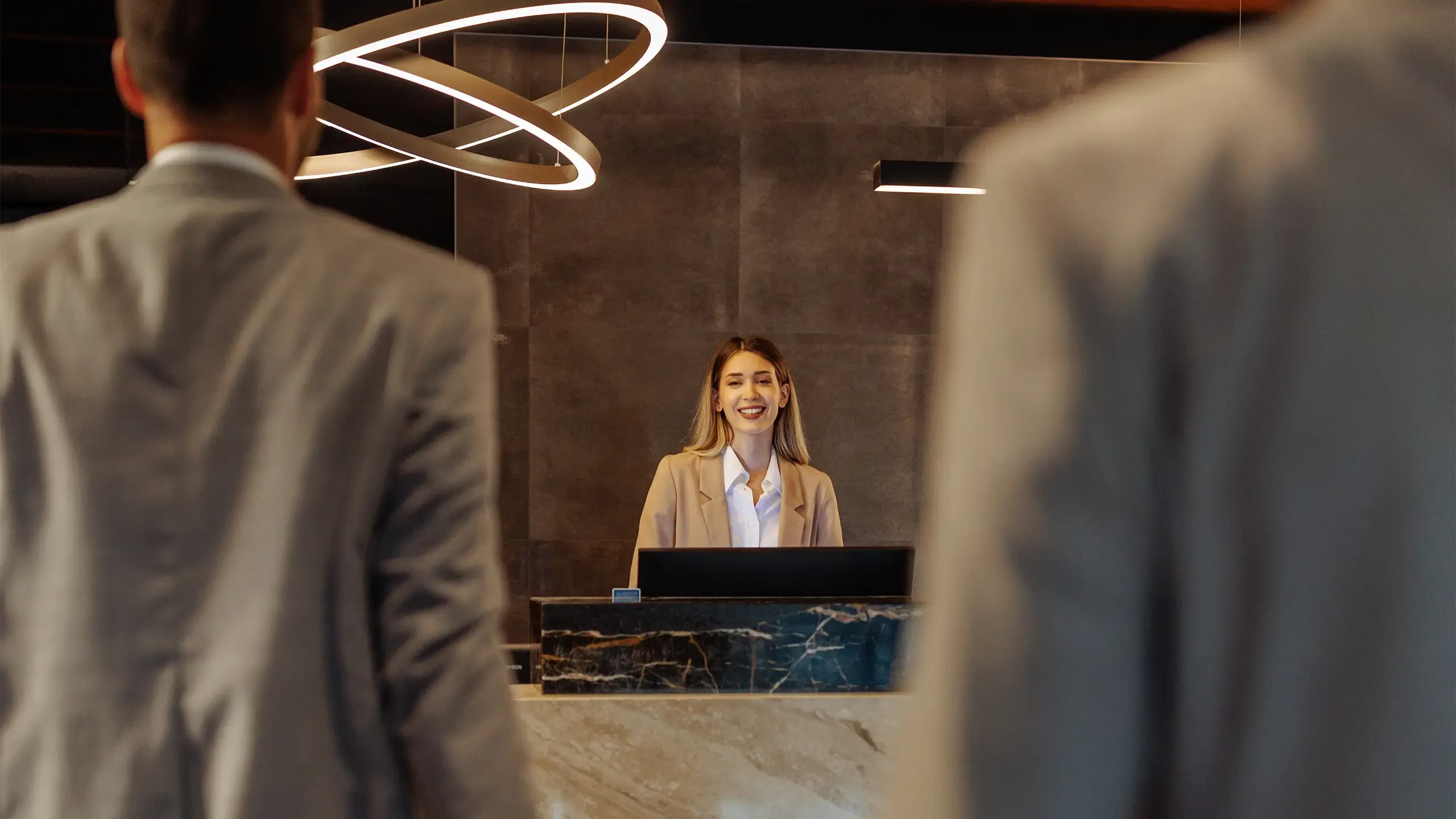 Two suited men walking in the hotel and being welcomed by a young Caucasian receptionist
