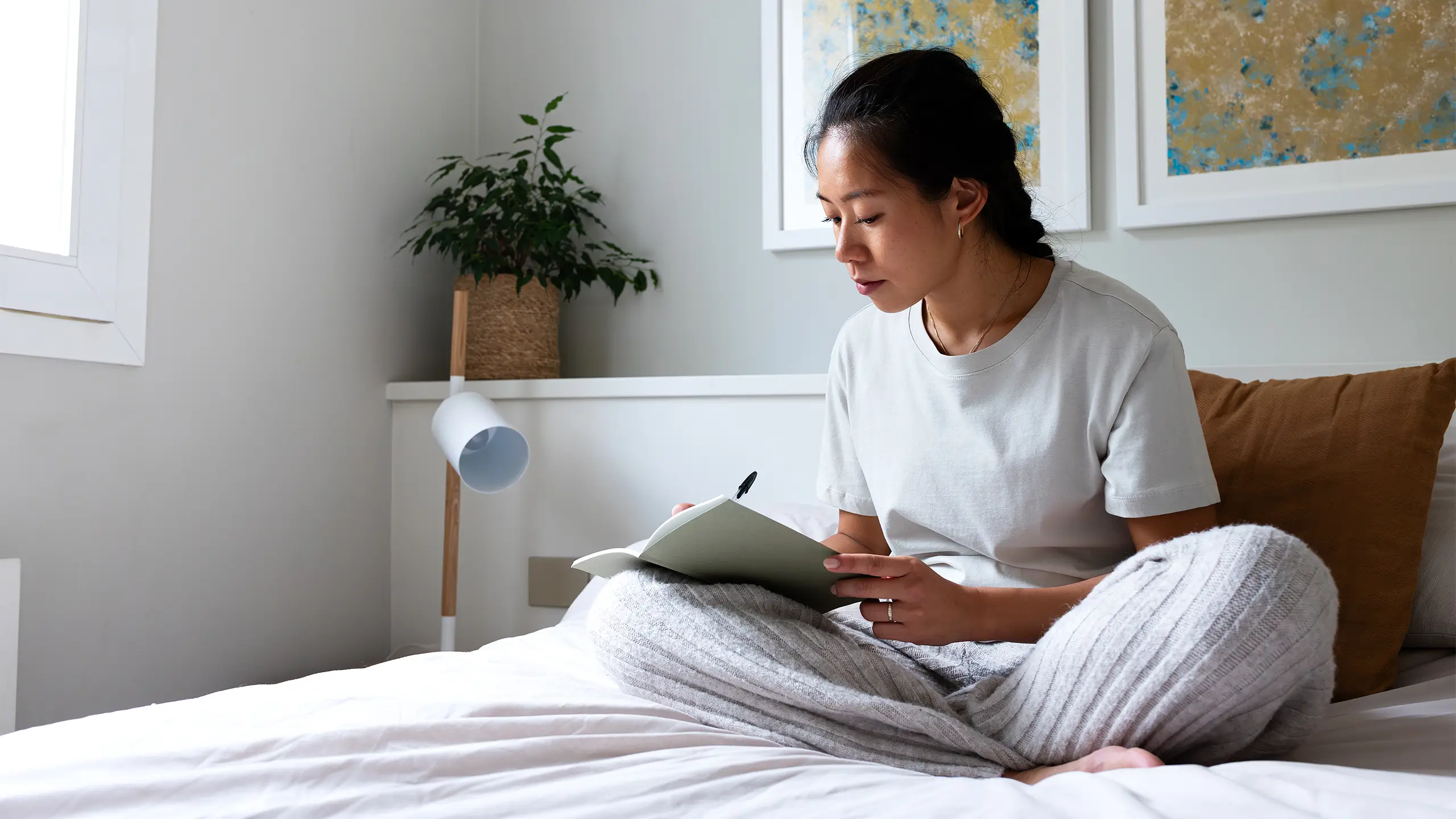 Pensive young Asian woman sitting on bed writing on journal in cozy bedroom. Copy space. Lifestyle concept.