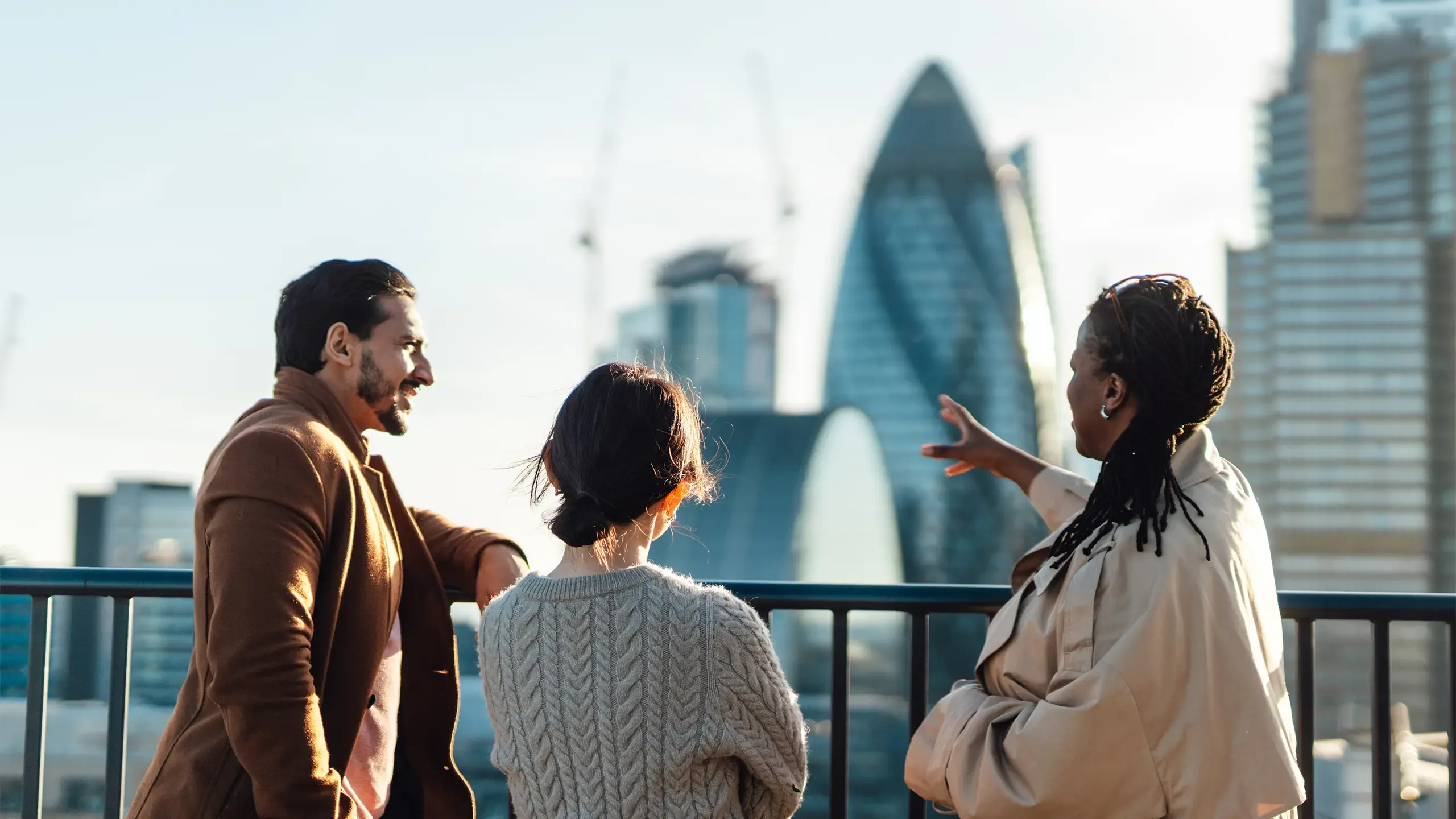 Multiracial business coworkers looking out across city at sunrise. Rear view of business people standing on rooftop in an office building against cityscape of London. Startup founders and partners looking to the bright future.