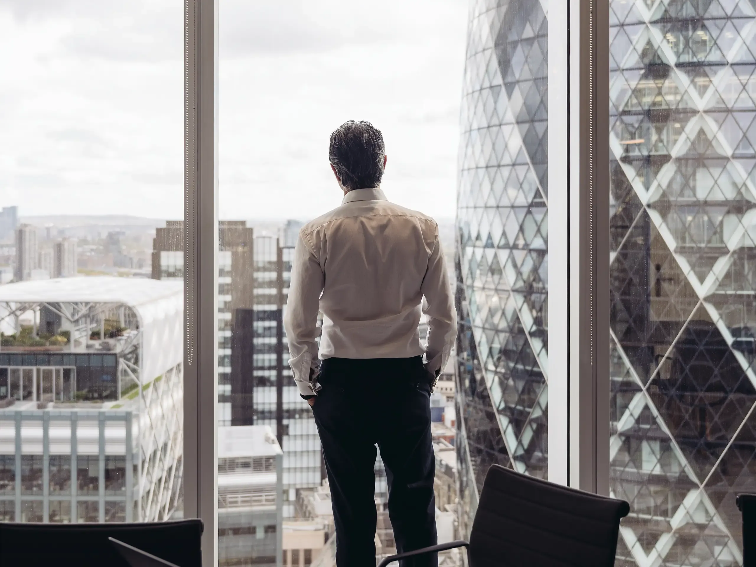 Rear view across conference table in modern board room of executive in early 40s standing with hands in pockets and looking at London’s financial district.