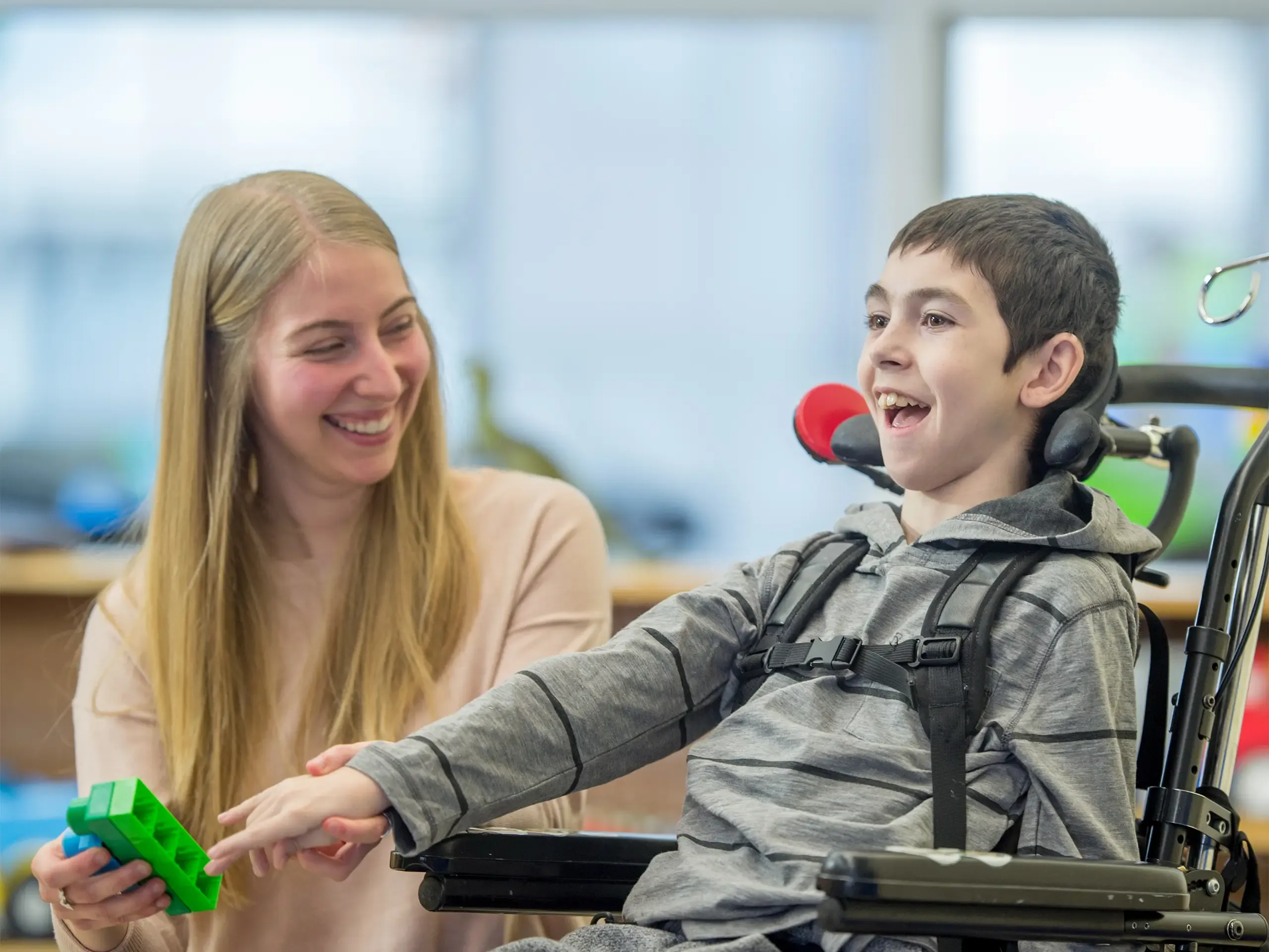 A boy in a wheelchair is indoors in his classroom. He is being assisted by his classroom helper. They are playing with colorful plastic blocks together.