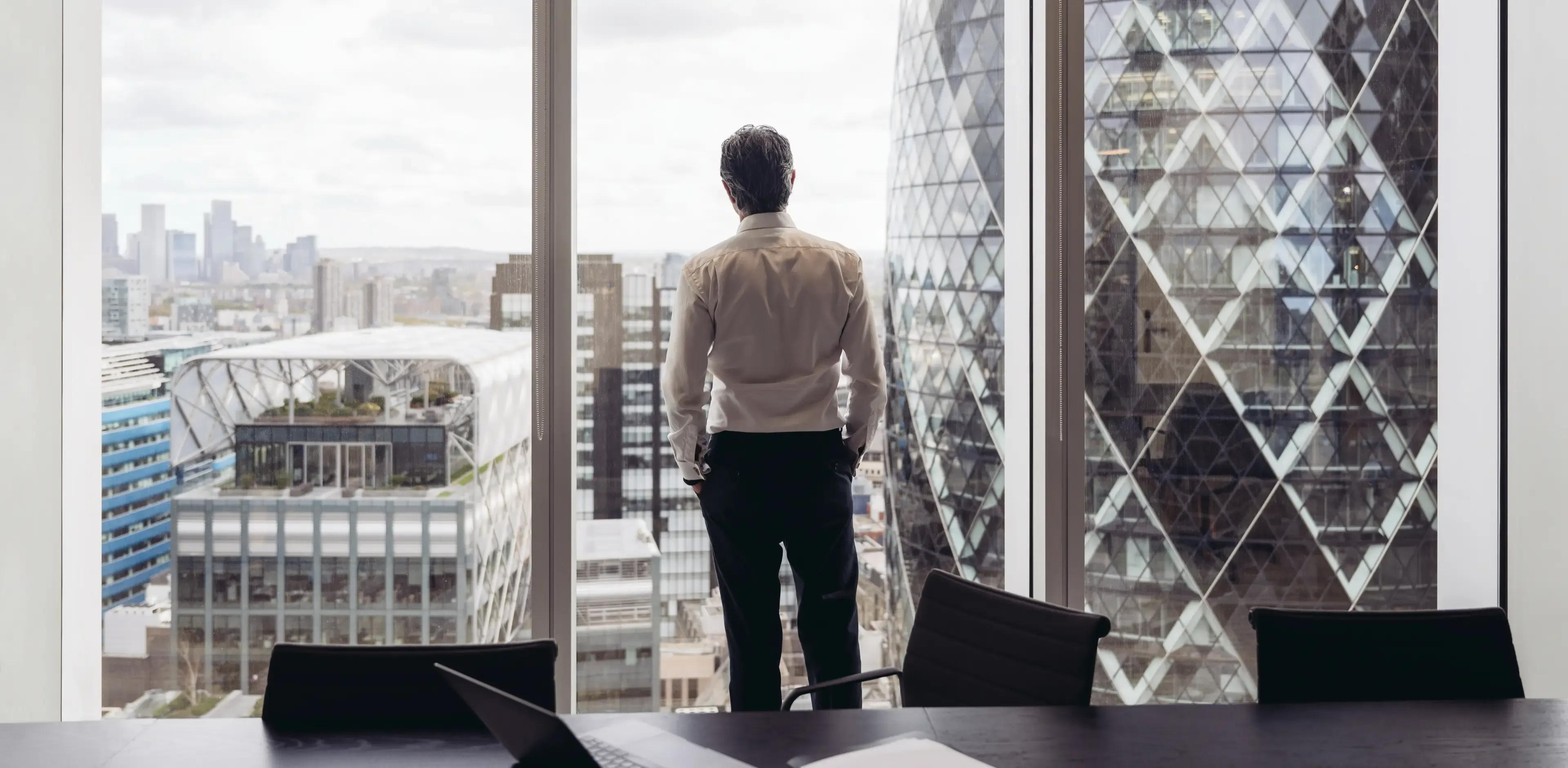 Rear view across conference table in modern board room of executive in early 40s standing with hands in pockets and looking at London’s financial district.